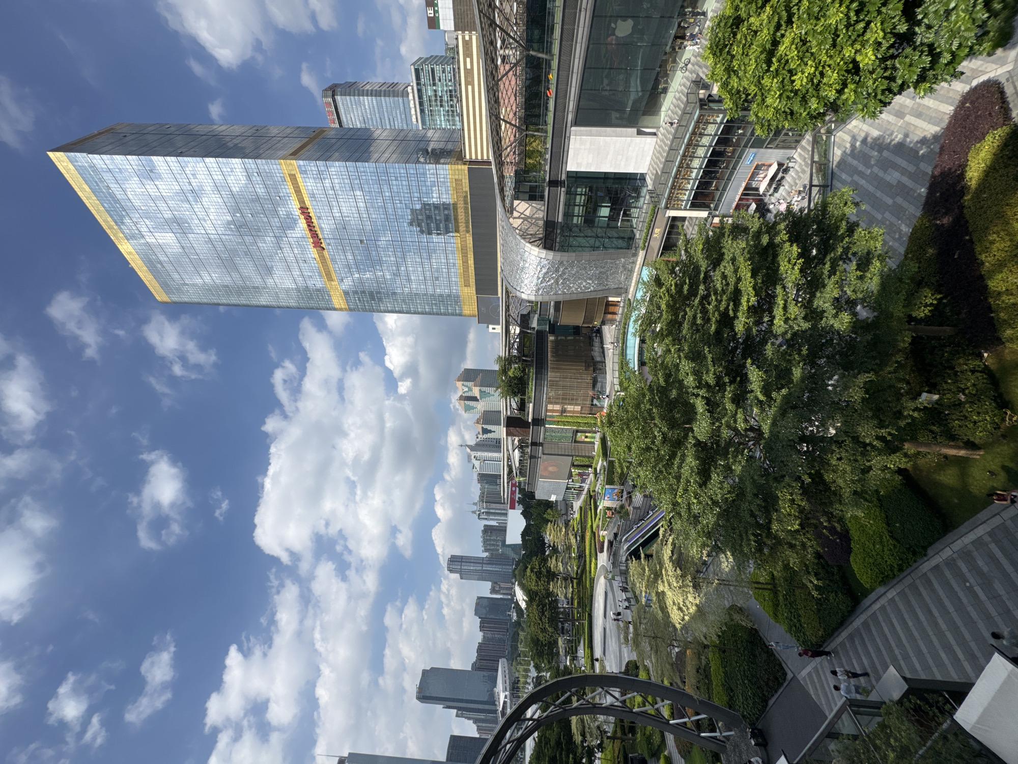 The image captures an urban landscape in Tianhe District, Guangzhou, China. The photograph is taken from an elevated position. To the left, a tall, rectangular, glass-covered skyscraper with horizontal yellow stripes and the word "Marriott" prominently displayed rises into a blue sky dotted with white clouds. Adjacent to this skyscraper is a modern building with a curved, silver exterior, possibly part of a shopping complex.
Below these structures, a meticulously maintained park or green space extends into the distance, featuring well-manicured trees, bushes, and walking paths. A variety of other skyscrapers and high-rise buildings are visible in the background, adding to the city's dense urban environment.
People are seen walking and interacting within the park area. Some are seated at outdoor cafes or restaurants, suggesting a leisure and commercial zone. A partial view of a metal structure in the foreground indicates the viewing location might be part of another building's architecture.
