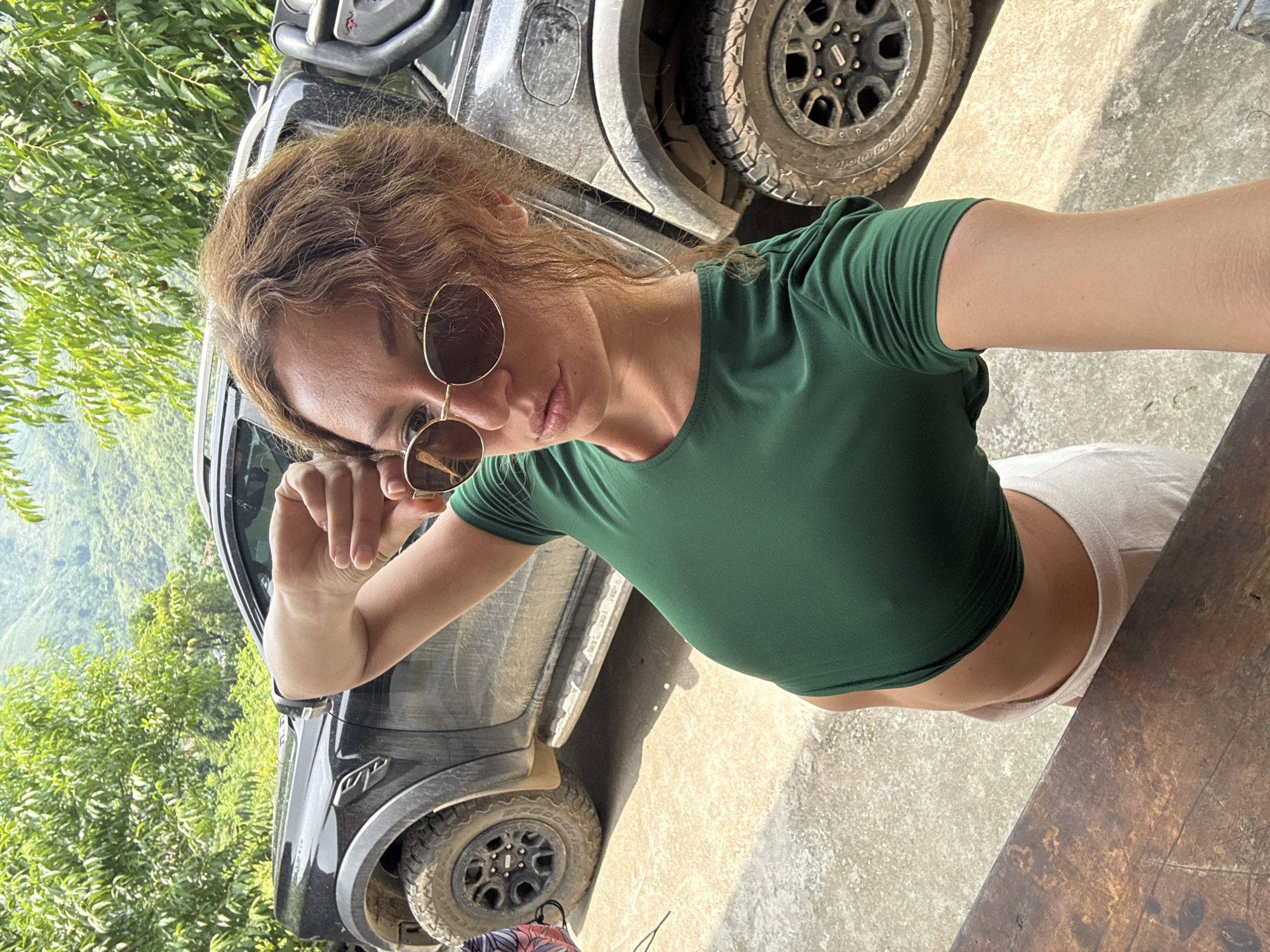 A selfie captures a woman posing in a verdant, mountainous landscape in Du Già Commune, Vietnam. The image is taken from a slightly high angle, looking down at the subject. The woman, with reddish-brown wavy hair, wears a dark green short-sleeved crop top and light-colored bottoms, with a bare midriff visible. She is wearing round, gold-rimmed sunglasses and looks directly at the camera. Her right hand is slightly raised, adjusting her sunglasses or holding the camera.
Behind her, a dark-colored SUV or truck, possibly a Ford Ranger Raptor, is visible, covered in mud on its tires and body, suggesting recent off-road travel. The letters "RAP" are discernible on the vehicle's side, and "GOO" (part of "Goodyear") is visible on the tire sidewall. Lush green trees and foliage dominate the background, indicating a dense, natural environment with a valley or mountain range extending into the distance. The ground immediately around the vehicle and extending into the foreground is a light-colored, possibly concrete or dirt, surface. A rustic wooden table or bench is present in the lower right foreground. The scene is brightly lit by what appears to be strong daylight, casting shadows.