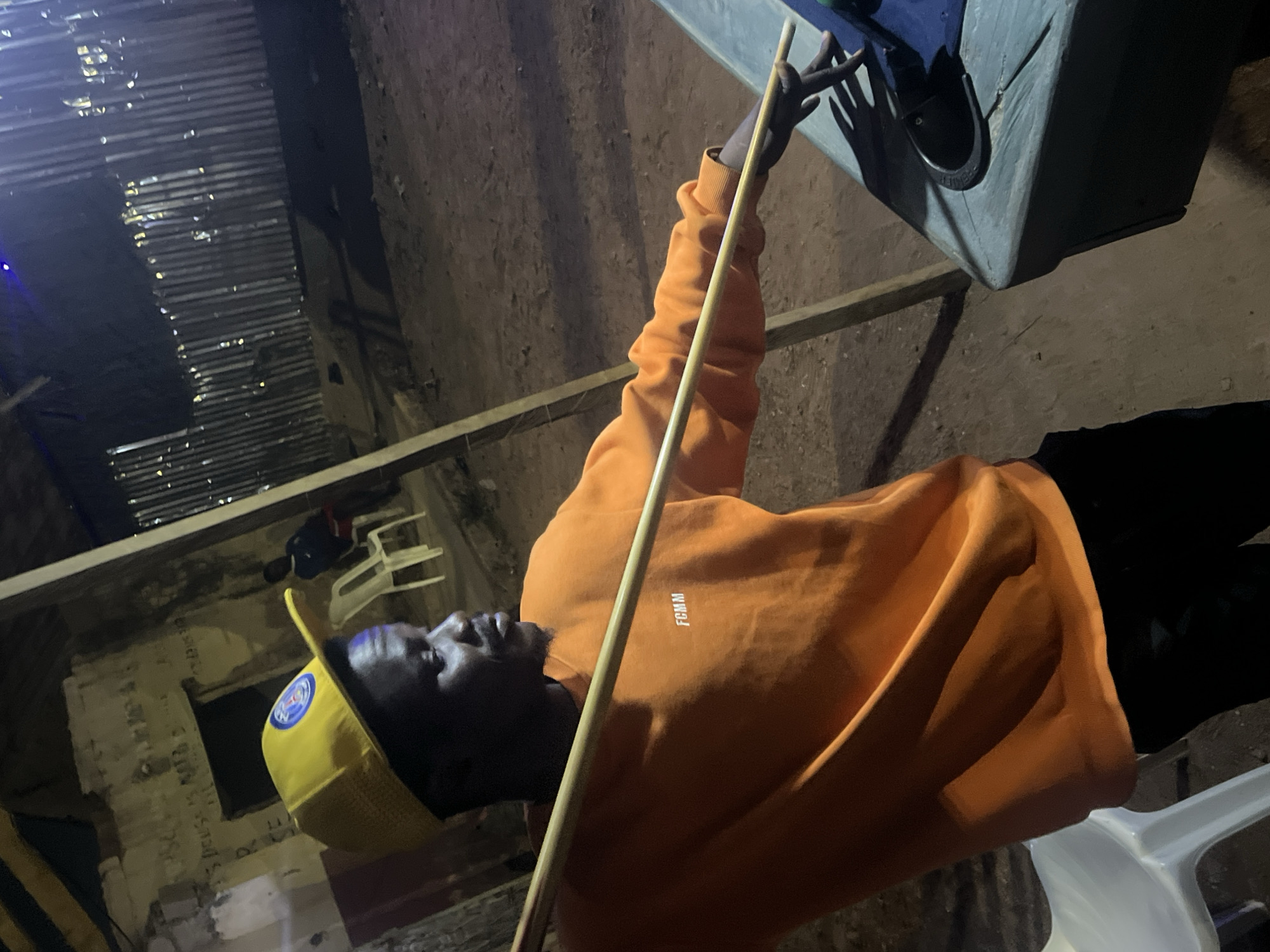 The image depicts a dark-skinned man in a dimly lit, rustic setting, possibly at night in Gura Top, Nigeria. The man, seen from the chest up, is wearing an orange hooded sweatshirt with "FCMM" written in white on the front, and a yellow baseball cap with an indiscernible circular logo. He has a beard and is looking upwards and to his left.
He holds a long, thin wooden stick or pole diagonally across his body with his left hand, which rests near a dark grey or blue rectangular object, possibly a speaker or cooler. In the background, to the man's left, there is a wall made of corrugated metal sheeting, reflecting some light. Further back, a white plastic chair and another dark object are visible. The ground appears to be rough dirt or concrete, traversed by a wooden beam. The overall environment suggests an informal or semi-enclosed space, like a shed, workshop, or casual gathering area.
The scene is lit by artificial light sources, casting shadows and highlighting certain areas, with a faint blue light visible on the far left. The man's expression is neutral and observant. On a distant wall in the background, faint graffiti or writing is visible, though mostly illegible, with some parts hinting at phrases like "...DRUGS IS NOT GOOD...".