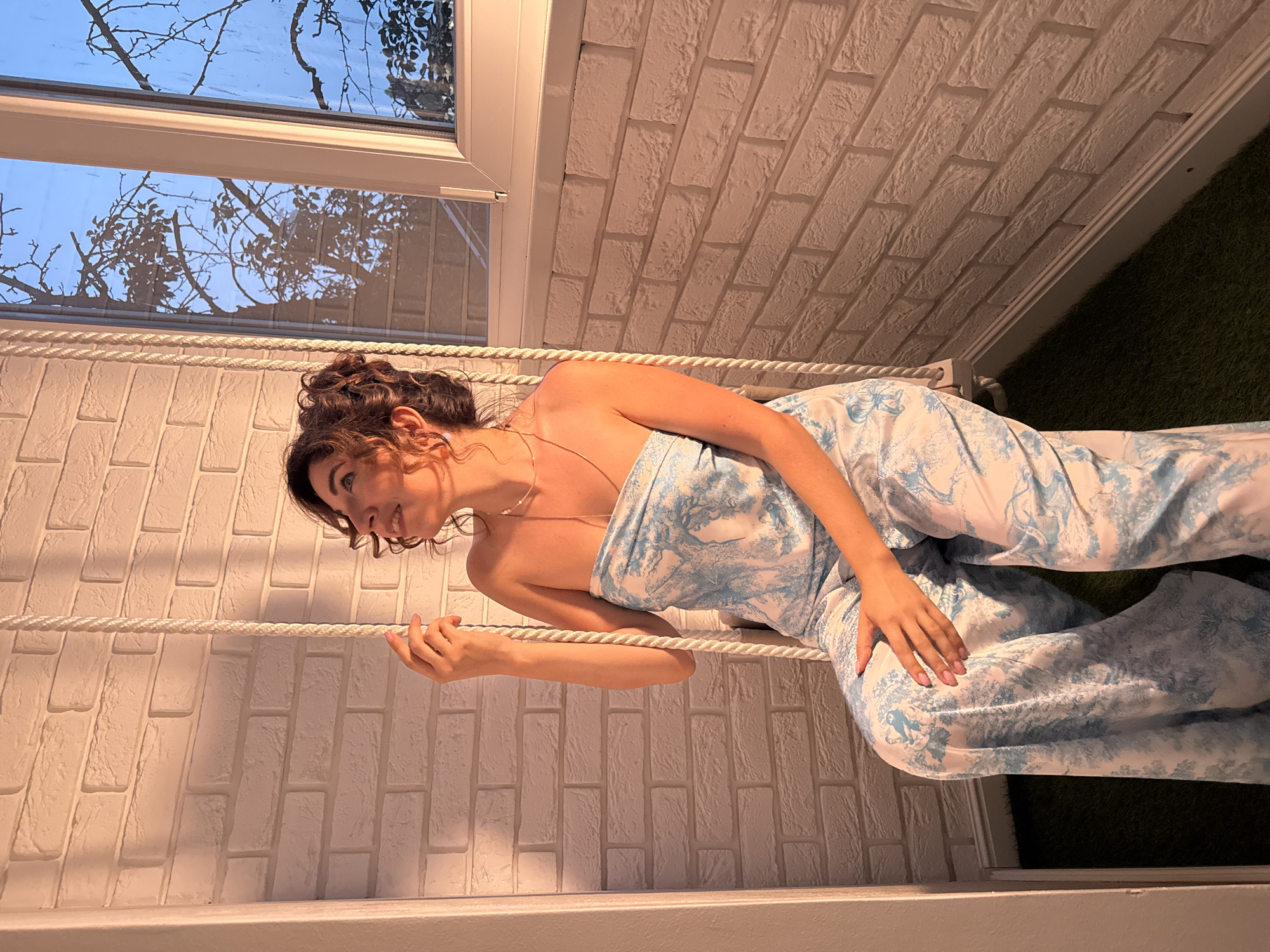 A woman with dark curly hair is posed on what appears to be a swing made of rope. She is wearing a strapless, wide-legged jumpsuit with a blue and white pattern. The background consists of a white brick wall and a window with a view of tree branches against a light blue sky, suggesting it's daytime. The woman is smiling and looking to her left, with her hand resting on the rope. The scene appears to be indoors, possibly a home or studio, given the controlled lighting and backdrop. The overall mood seems relaxed and cheerful. There is no visible text in the image.