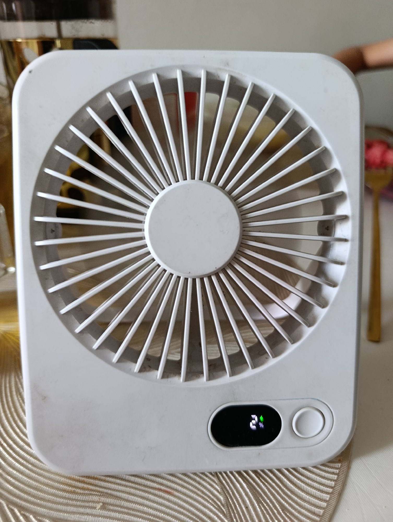 A white, square-shaped portable fan is positioned on a beige, textured surface. The fan has a circular grill with radiating lines and a central circular button. On the lower right side of the fan, there is a small black digital display showing "24%" and a green leaf icon, indicating some form of environmental reading, possibly temperature or humidity. Next to the display is a circular white button, likely for power or control. The background is blurred, but suggests an indoor setting with some objects visible, including what appears to be a glass container with a golden rim on the left and a gold utensil on the right. The overall scene is clean and well-lit.