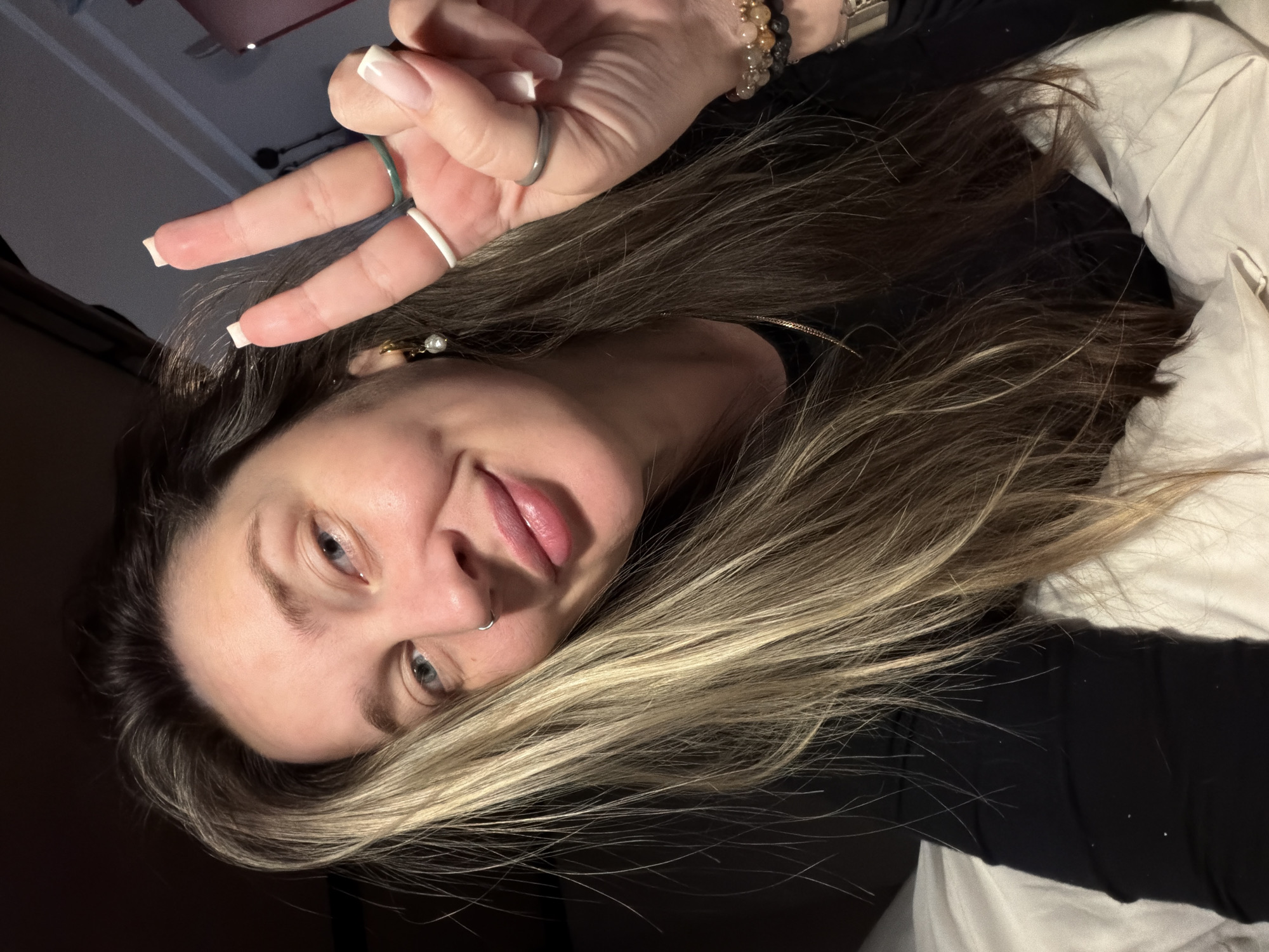 A medium close-up shot, angled diagonally, features a woman with long, layered brown hair highlighted with blonde streaks. She is looking directly at the camera with a subtle smile and her head slightly tilted. Her fair skin, blue eyes, and full lips are visible, along with a small silver hoop nose ring.
Her right hand is raised, making a peace sign towards the upper left of the frame. Her fingers display neat, square-shaped nails with white French tips. Several rings adorn her fingers: a thin green band on her index finger, a white band on her middle finger, and a silver ring on her ring finger. Her wrist is adorned with multiple beaded bracelets, some appearing to be made of dark, textured stones (possibly lava stone) and lighter, smooth beads.
She is wearing a black top or sweater, and a small pearl earring is visible on her right ear. A white pillow or bedding is visible in the lower right and upper right background, suggesting an indoor setting, likely a bedroom. The lighting appears soft and artificial, hinting at an evening or nighttime shot. The overall mood is relaxed and friendly. There is no visible text in the image. The location context is Moscow, Russia.