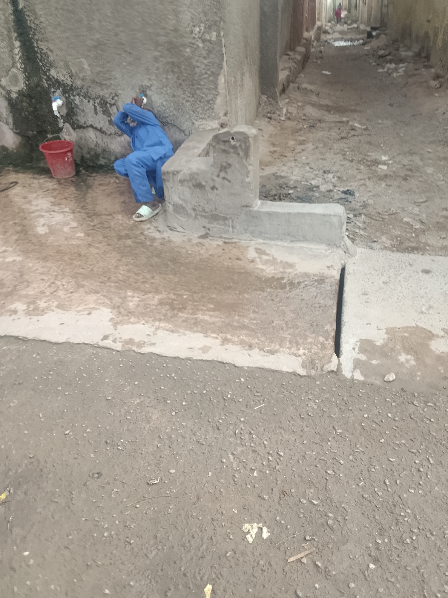 A man in blue clothing washes his face at a public tap in an alleyway in Pantame, Nigeria. The tap is connected to a weathered concrete structure. A red bucket sits beneath the tap. The ground is wet around the tap and runoff flows into a drain. A person in red walks down the alleyway. The environment appears impoverished.