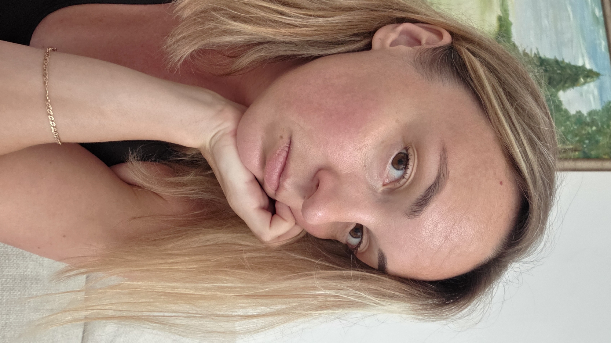The image shows a close-up, sideways portrait of a young woman with blonde hair. She is lying down, with her head resting on her arm and hand. Her eyes are open and looking towards the camera. She has light-colored skin and her lips are slightly parted. There is a subtle blush on her cheeks. A thin, gold chain bracelet is visible on her arm. The background is blurred, but appears to be an indoor setting with a painting on the wall that depicts a landscape with trees. The lighting suggests it could be daytime. The woman's pose and expression could be interpreted as contemplative or relaxed. The location is identified as Guía de Isora, Spain.