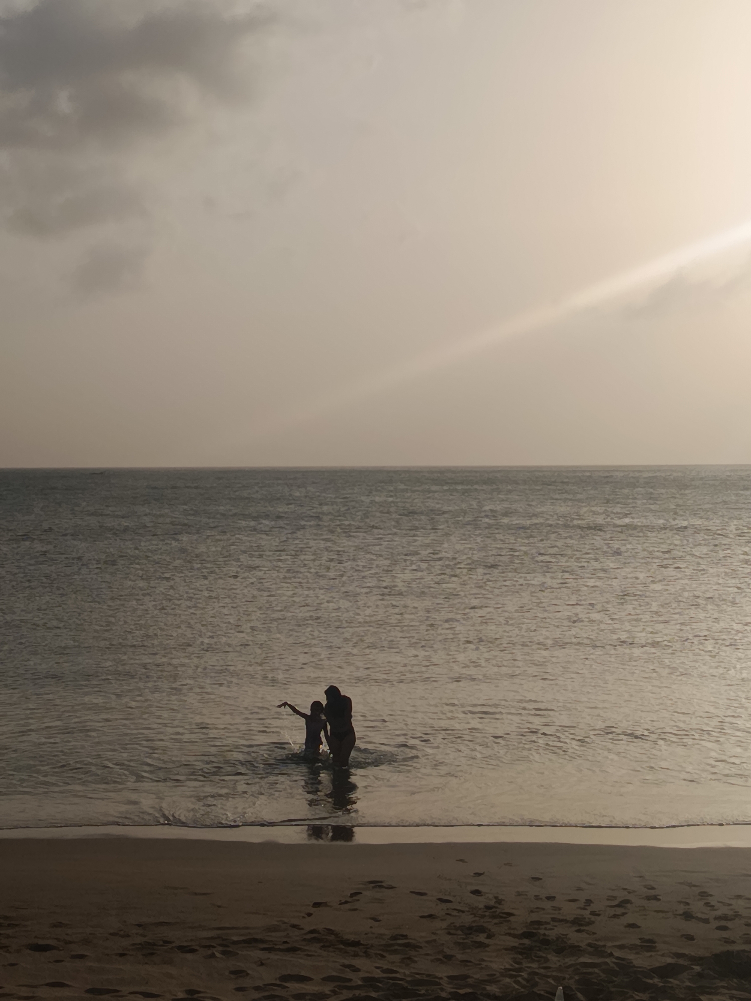 Two people are standing in shallow water at the beach in Juan Griego, Venezuela. The sky is overcast with a ray of light shining through. The beach is sandy with several footprints. The water is calm.