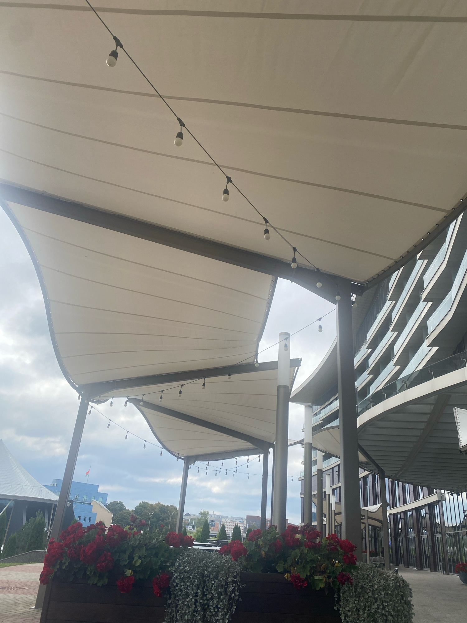The image captures an outdoor area with a modern architectural design, likely a cafe or restaurant terrace, located in Minsk, Belarus. In the foreground, planters filled with vibrant red flowers and trailing green foliage are visible. Above, a series of white fabric awnings, supported by dark metal beams and columns, extend over the seating area. String lights with bare bulbs hang between the awnings, adding a decorative touch.
In the background, a contemporary building with a curved facade and multiple levels of windows with diagonal dividers is prominent. Further in the distance, a building with a conical roof and a flag on top can be seen, along with other city structures and trees under a cloudy sky. The overall scene suggests a daytime setting, possibly a slightly overcast day.
