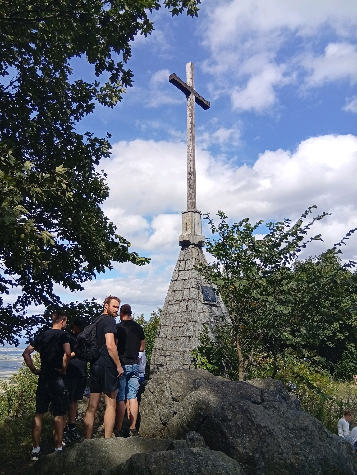 The image shows a group of individuals standing on a rocky outcrop, with a large wooden cross on a stone pedestal in the background. The cross is positioned against a bright blue sky with scattered clouds. The surrounding environment is natural, with trees and foliage present. The people are dressed casually in t-shirts and shorts. One individual, in the foreground, is facing away from the camera and appears to be looking at the cross. Another person is partially visible to the right, also looking in the same direction. The stone pedestal of the cross has a plaque affixed to it. The scene suggests a visit to a scenic or historical location.
The location is likely the summit of Mount Ślęża near Sobótka, Poland, a popular pilgrimage and tourist site featuring a prominent cross and a historical monument.