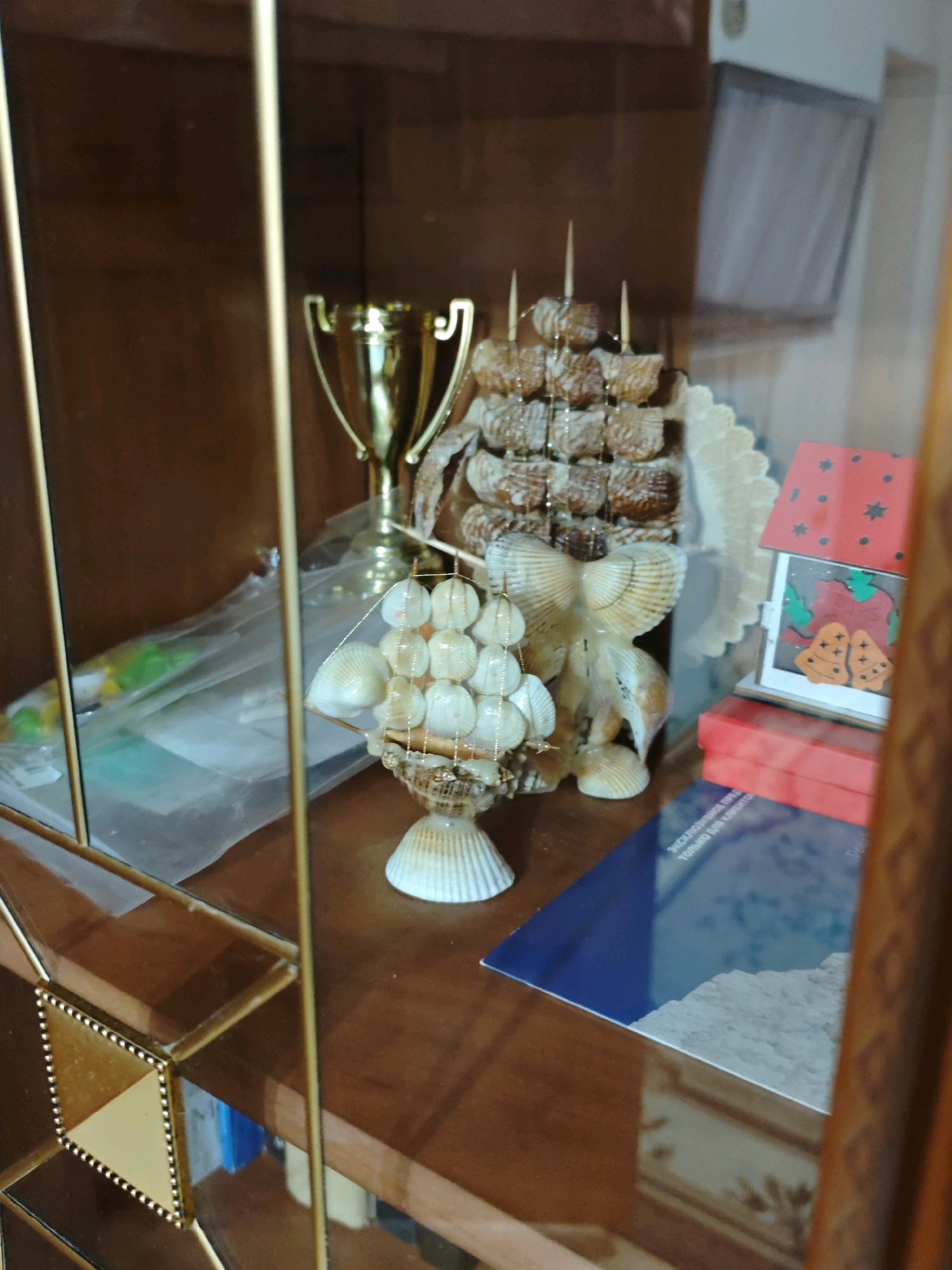 A framed photograph inside a display cabinet shows a golden trophy, a seashell ship, and a small red house decoration with bells. The cabinet has wooden shelves and glass doors. The photo's reflection shows a tiled wall with floral patterns. This image was taken in Yevpatoriya, Ukraine.