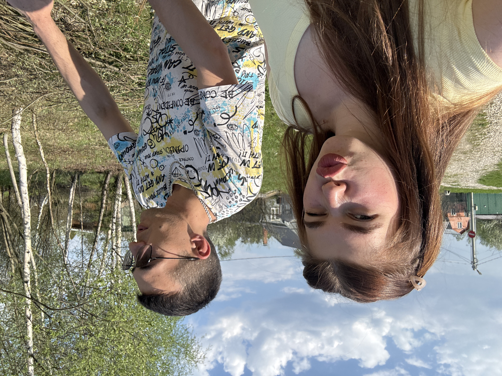 A photo taken in Шевлягино, Russia shows two people in an outdoor setting. The image appears to be rotated 180 degrees. On the left is a male wearing glasses and a printed shirt, standing near birch trees. On the right, a female with long brown hair is wearing a yellow top. The background includes houses, a blue sky, and some clouds.