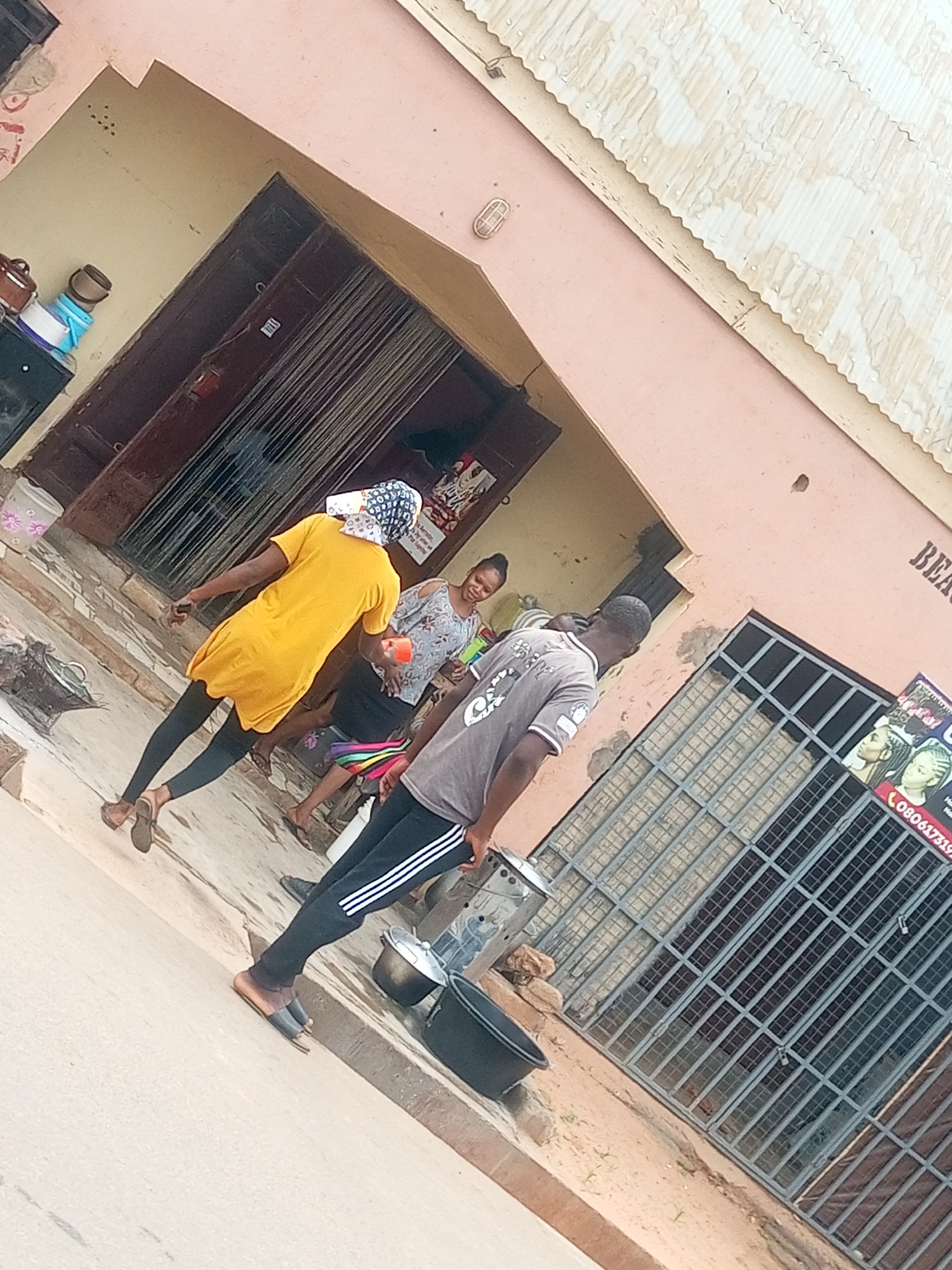 In Gombe, Nigeria, a scene unfolds near a building with a pink facade. Three individuals are present. One person, seen from the back, wears a yellow dress and dark leggings. Another, a woman in a patterned shirt, stands with her back to the viewer, holding a red cup. A third person, a man, is seen from the side, wearing a gray shirt with white stripes on the sleeves and dark pants. The ground is paved, and to the right, a window with metal bars is visible. Signs displaying images of hairstyles and a phone number are attached to the window. Two buckets are placed on the ground.