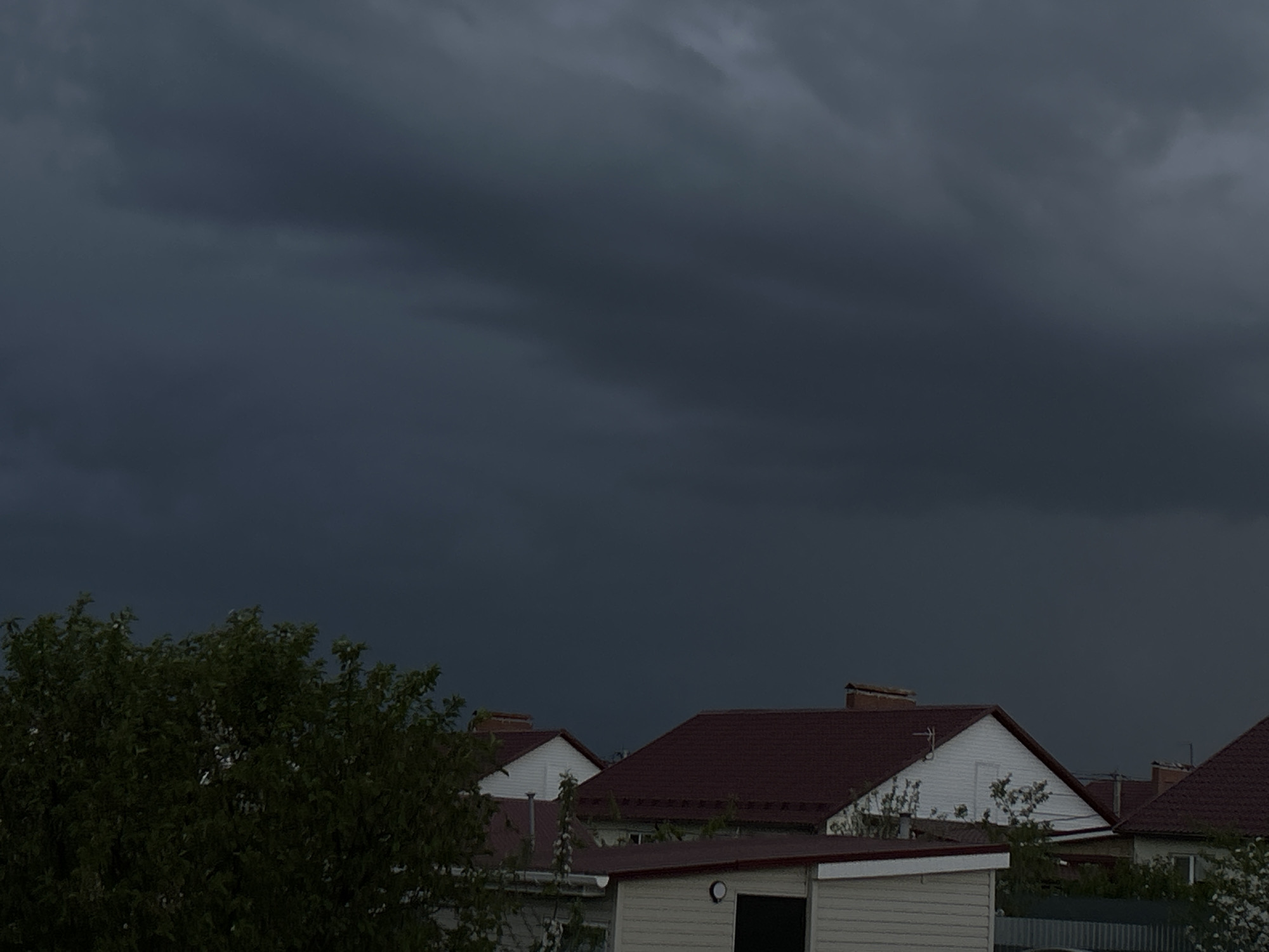 The image depicts a residential area in Узюково, Russia, under a dark, ominous sky suggesting an approaching storm. Several houses with red tile roofs and white siding are visible. In the foreground, a leafy tree obscures part of the scene. A small outbuilding or shed is also visible with a dark-colored door. There are no people visible in the image. The overall environment is dark and overcast.