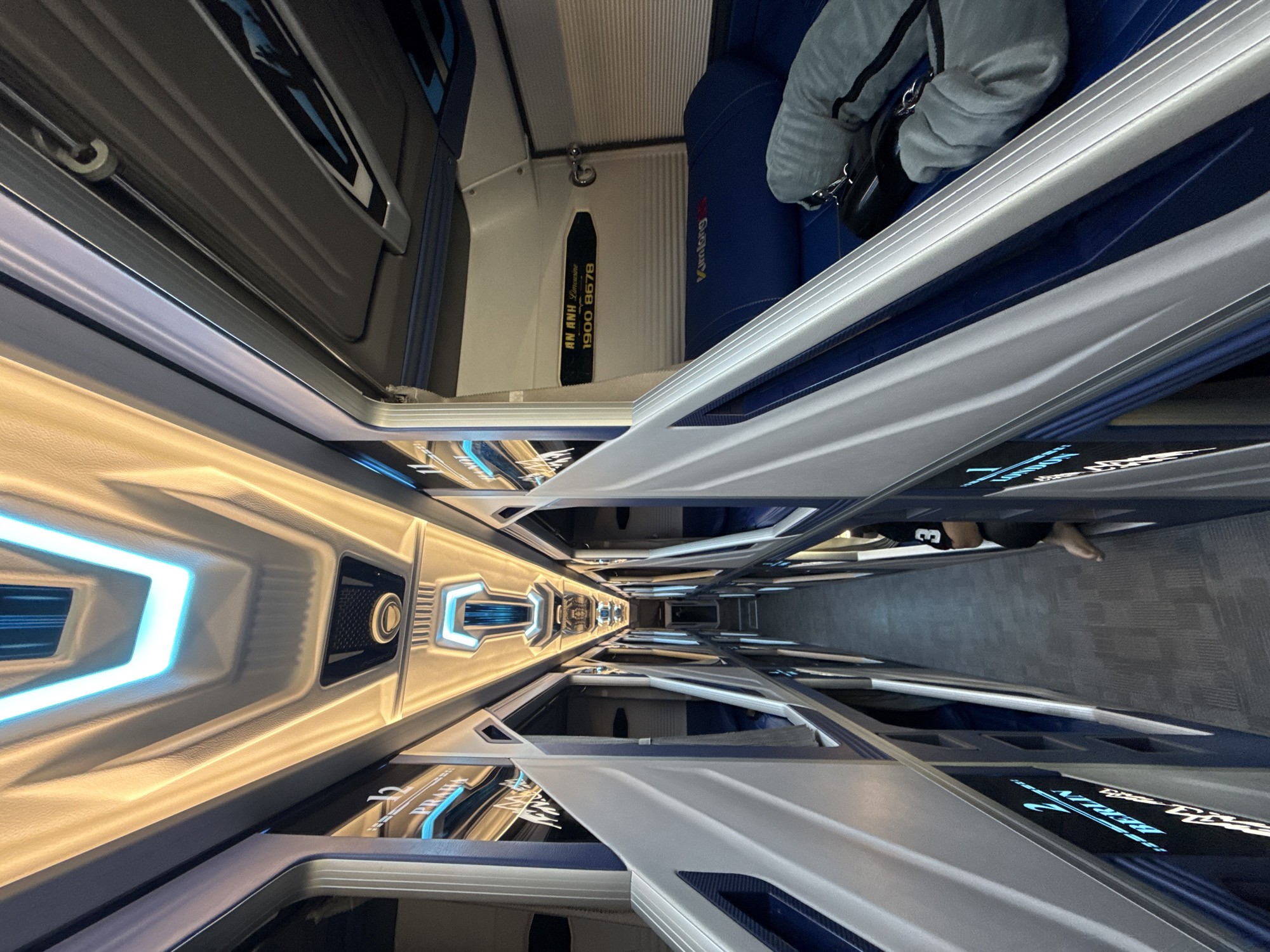 The image shows the interior of a sleeper bus, likely a limousine bus service, within the city of Da Lat, Vietnam. The bus features individual sleeping pods arranged along a central aisle. The pods are lined with blue upholstery and have illuminated signs indicating destinations like "Berlin" and "London" above them. The ceiling of the bus is adorned with dramatic, futuristic lighting, combining warm yellow and cool blue tones, creating a modern and luxurious ambiance. In one of the visible pods, a person is partially visible, with a grey blanket and what appears to be a bag. The floor of the aisle is carpeted in a grey, textured material. The overall scene depicts a well-appointed interior designed for long-distance travel comfort.