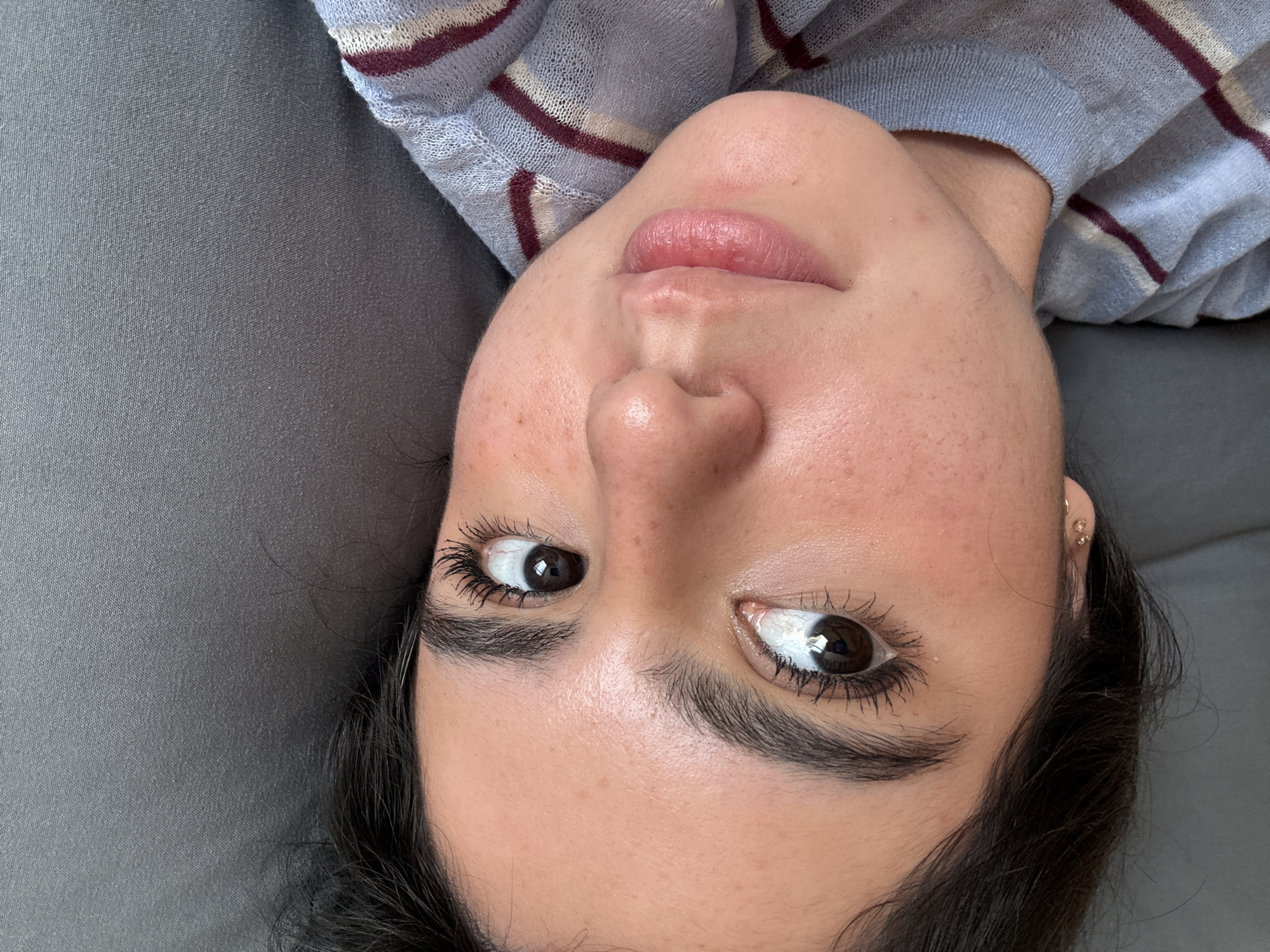 A close-up, eye-level shot of a woman's face, angled upside down. She has dark hair, brown eyes, and fair skin with visible pores and freckles. Her lips are full and slightly parted. She is wearing mascara and small earrings. She is lying on a gray surface, possibly a couch or bed, wearing a light blue and maroon striped sweater. There is no information to suggest that the image was taken in San Jerónimo Chicahualco, Mexico.
