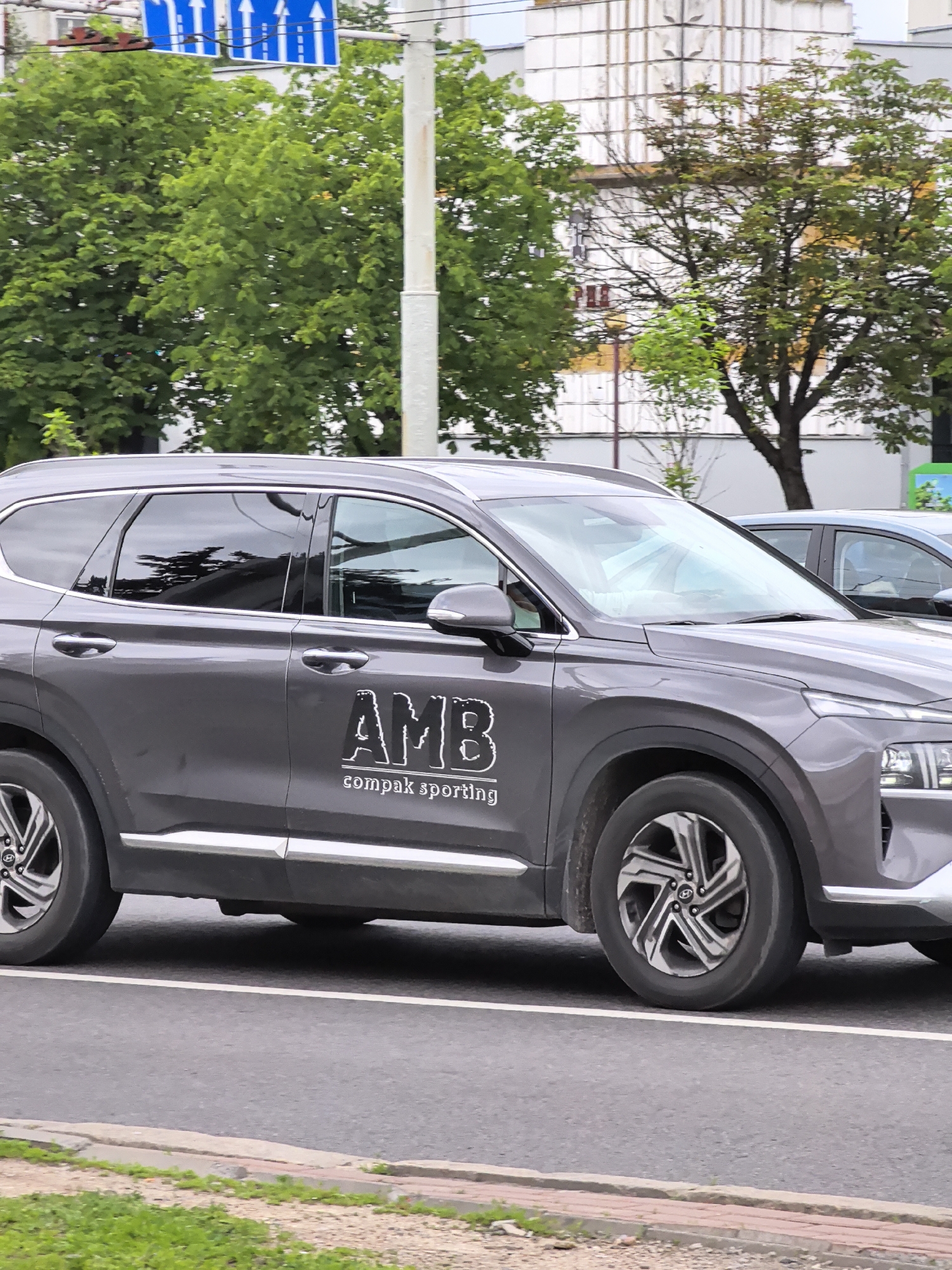 A dark gray SUV is parked on a city street. The SUV has tinted windows and a chrome trim line along the bottom of the doors. On the side of the SUV, there is a logo that says "AMB" in large, distressed white letters, with smaller text below that reads "compak sporting". The SUV is on the right side of the frame. In the background, there are green trees, a white building with some architectural details, and a utility pole. A blue traffic sign with white arrows is visible in the upper left corner. The street is paved with asphalt, and there is a white line marking the lane. A grassy area with some gravel is visible in the foreground.