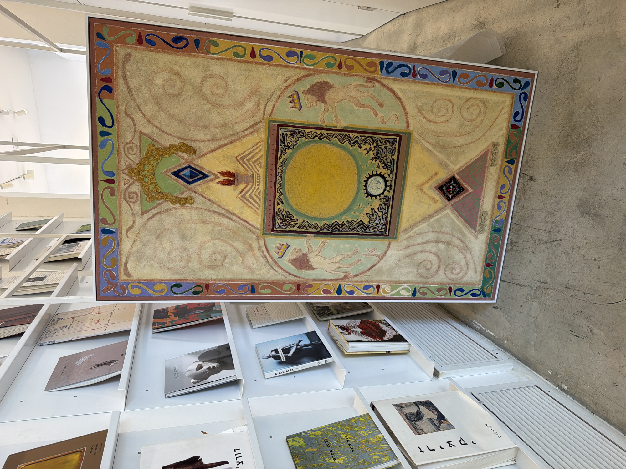 The image shows a large, framed artwork displayed in a brightly lit, minimalist gallery space. The artwork, with a pale yellow background and intricate borders, features a central circular motif surrounded by decorative elements and stylized figures, including lions with crowns. The gallery shelves on the left are filled with books, some of which are visible with their titles and covers. The overall impression is one of a clean, modern exhibition space, likely in Tel Aviv, Israel, given the presence of Hebrew text on some book spines and the general aesthetic often found in Tel Aviv galleries.