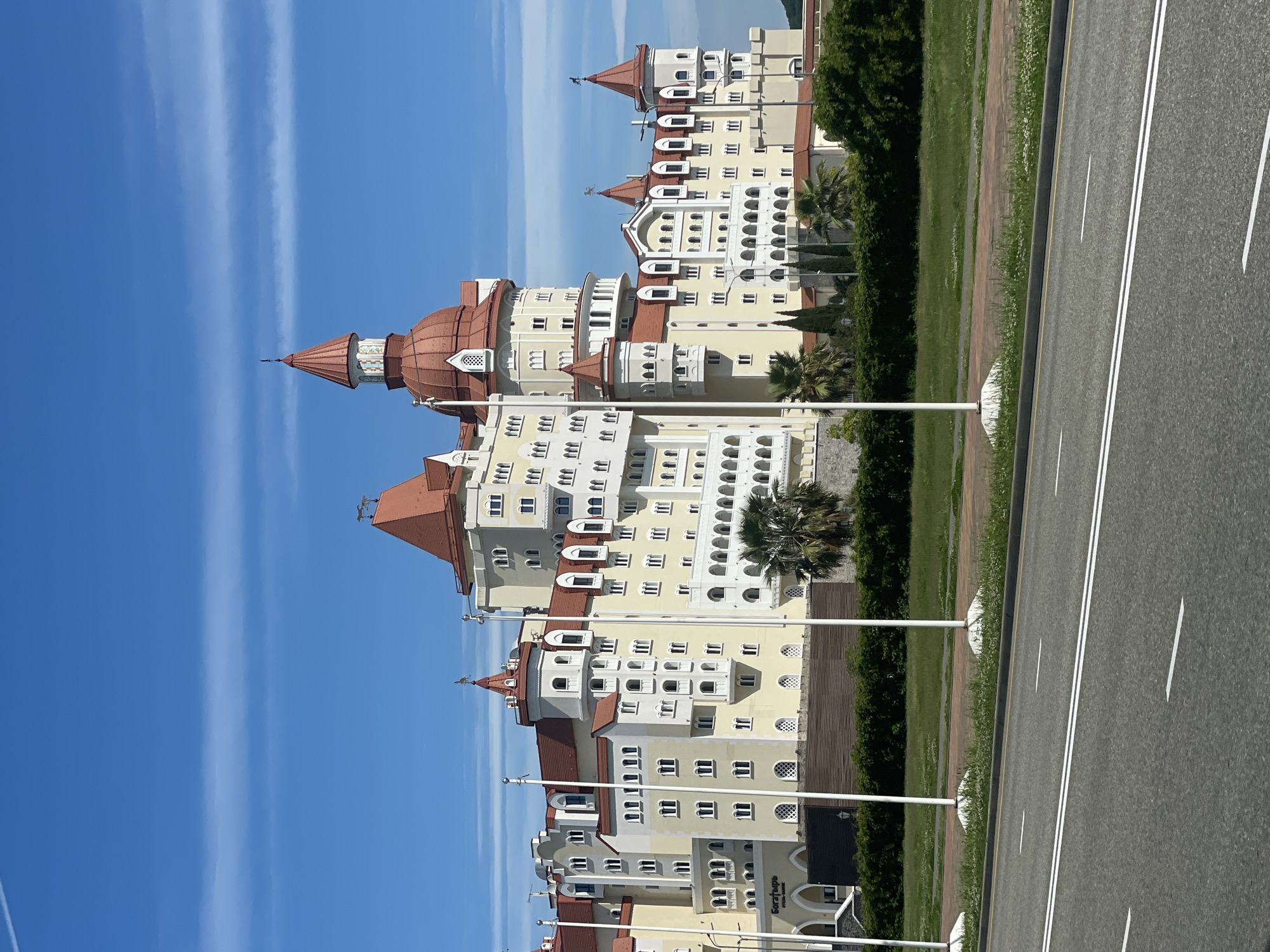 The image depicts a multi-story building with a unique architectural style, featuring cream-colored walls, red roofing, and numerous windows. The building is positioned next to a road with double white lines marking lanes. A strip of green foliage separates the building from the road. Above, a clear blue sky stretches with thin white clouds. The location is Sirius, Russia.