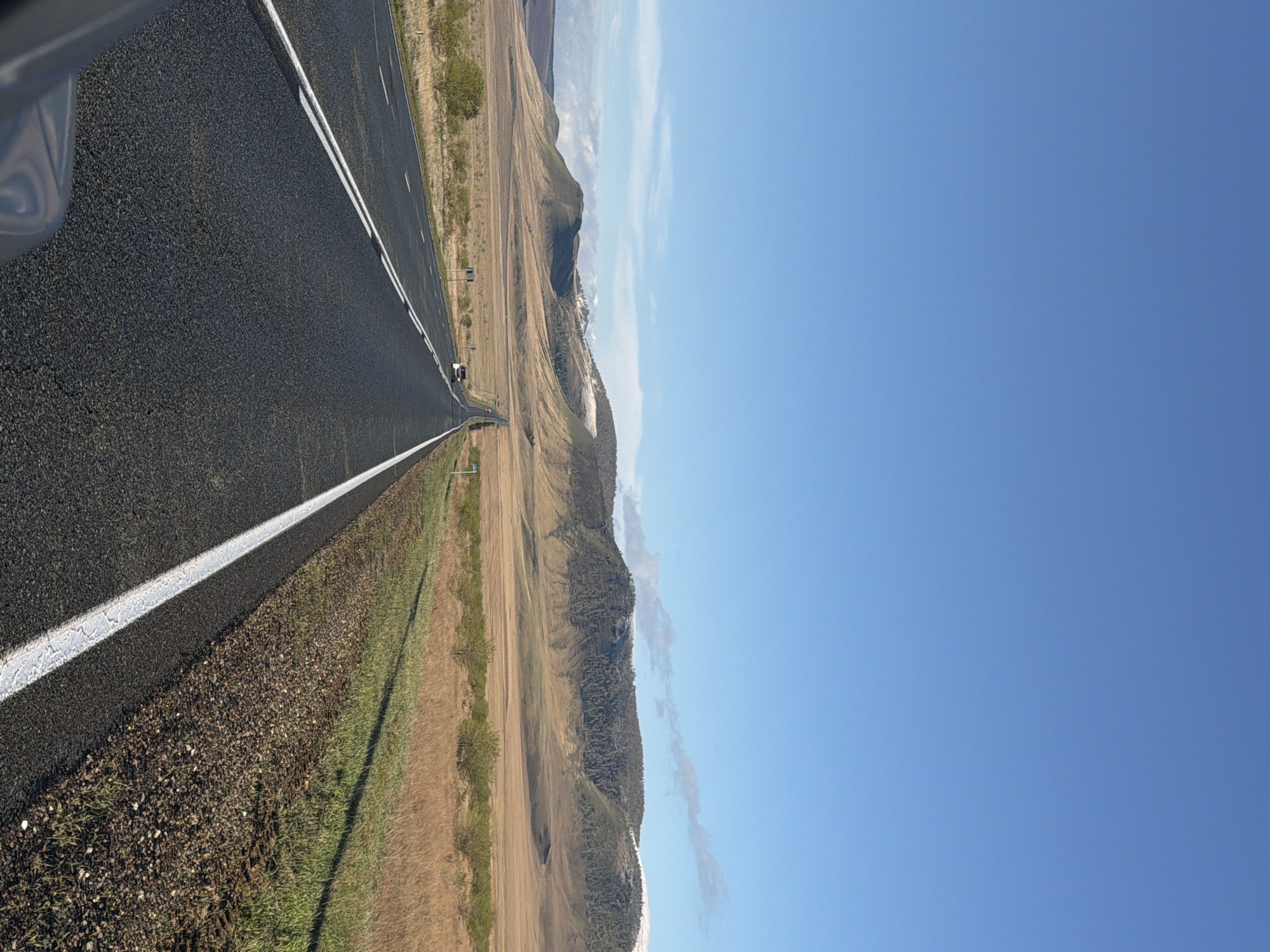 A road stretches horizontally across the left side of the image, marked with a white line. A car's side panel is visible in the upper-left corner. To the right of the road, a grassy shoulder gives way to arid fields and rolling hills. A clear, blue sky dominates the right side of the frame. A location in the city of Bazandaikha, Russia, is not possible to verify from the visual data provided.
