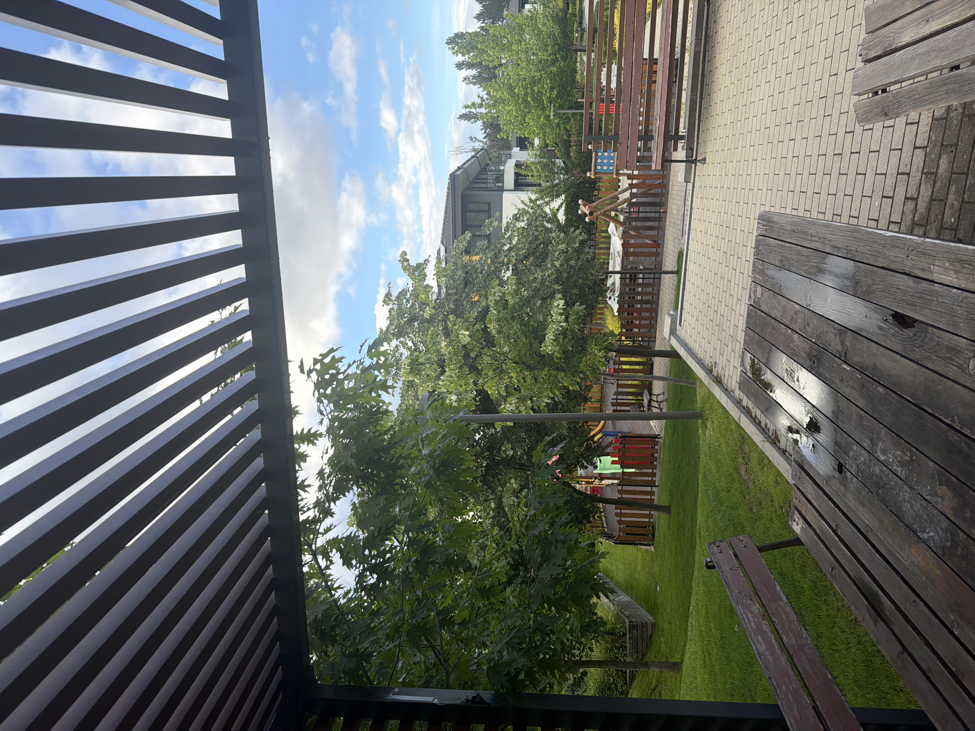 This landscape-oriented image captures a serene outdoor setting, likely a park or communal garden within a residential area. In the immediate foreground on the left, a dark slatted structure, possibly part of a pergola or trellis, partially frames the scene, casting distinct shadows. To the right, two wooden benches are visible; a larger, rustic picnic-style table or bench and a smaller, darker bench, both showing signs of weathering.
The midground features a light-colored paved pathway made of interlocking bricks, stretching towards the background. Adjacent to the paving is a vibrant green lawn area, dotted with several trees boasting lush green foliage, suggesting a warm season. A children's playground is discernible among the trees, featuring wooden structures and colorful elements in red, green, and blue.
In the background, a modern building with dark grey and white facades stands, partially obscured by trees. The sky above is a clear blue with scattered white, fluffy clouds. The scene is devoid of people, contributing to a calm and peaceful atmosphere. The bright lighting, visible shadows from the slatted structure and benches, and clear sky indicate it is daytime, likely morning or afternoon. While no specific activity is occurring, the setup suggests a space for relaxation, play, and community gathering. No visible text is present in the image. The location context is Sofia, Bulgaria.