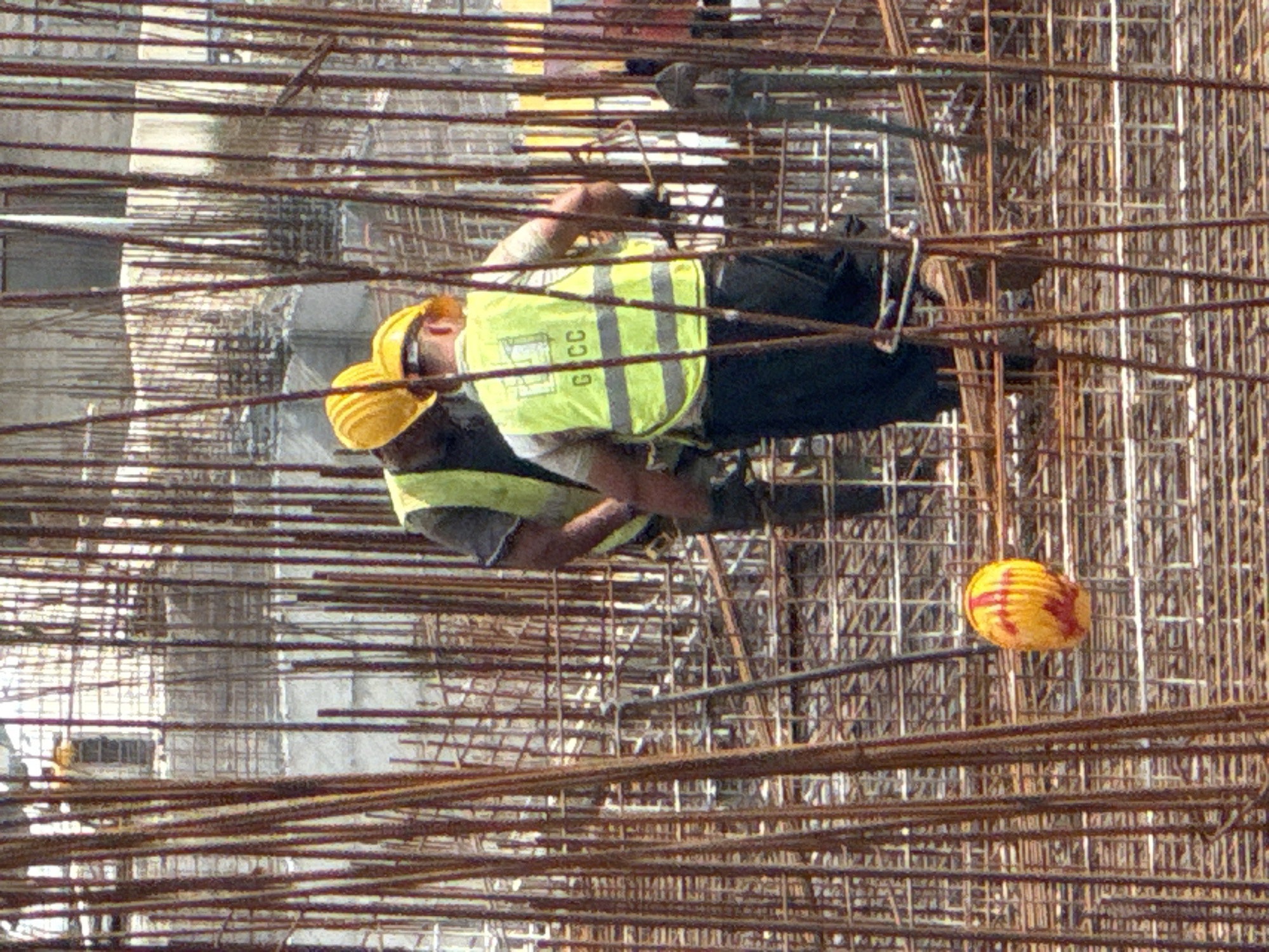 The image depicts a construction site in Boreti, Montenegro. Two construction workers are visible, both wearing yellow hard hats and high-visibility vests. One worker, on the left, is wearing a yellow vest with gray reflective stripes and appears to be working with rebar. They are also wearing dark pants and a white undershirt. The second worker, to the right, is also wearing a yellow hard hat and a yellow vest with gray reflective stripes over dark clothing.
The scene is dominated by a dense network of rusty rebar, indicative of concrete reinforcement work for a large structure, likely a building. The lighting suggests it is daytime, with sunlight casting shadows, but the specific time of day is not discernible. The weather appears to be clear.
A notable object in the image is a yellow and red striped ball, possibly a safety marker or a child's toy, suspended among the rebar structure in the mid-ground to the right. The overall impression is one of ongoing construction activity, with an emphasis on the raw materials and the labor involved. The crops provided offer closer views of the workers and their immediate surroundings, highlighting the details of their gear and the intricate rebar framework. There is no discernible text in the image other than what may be on the workers' vests, which appear to have lettering that could be "GCCC".