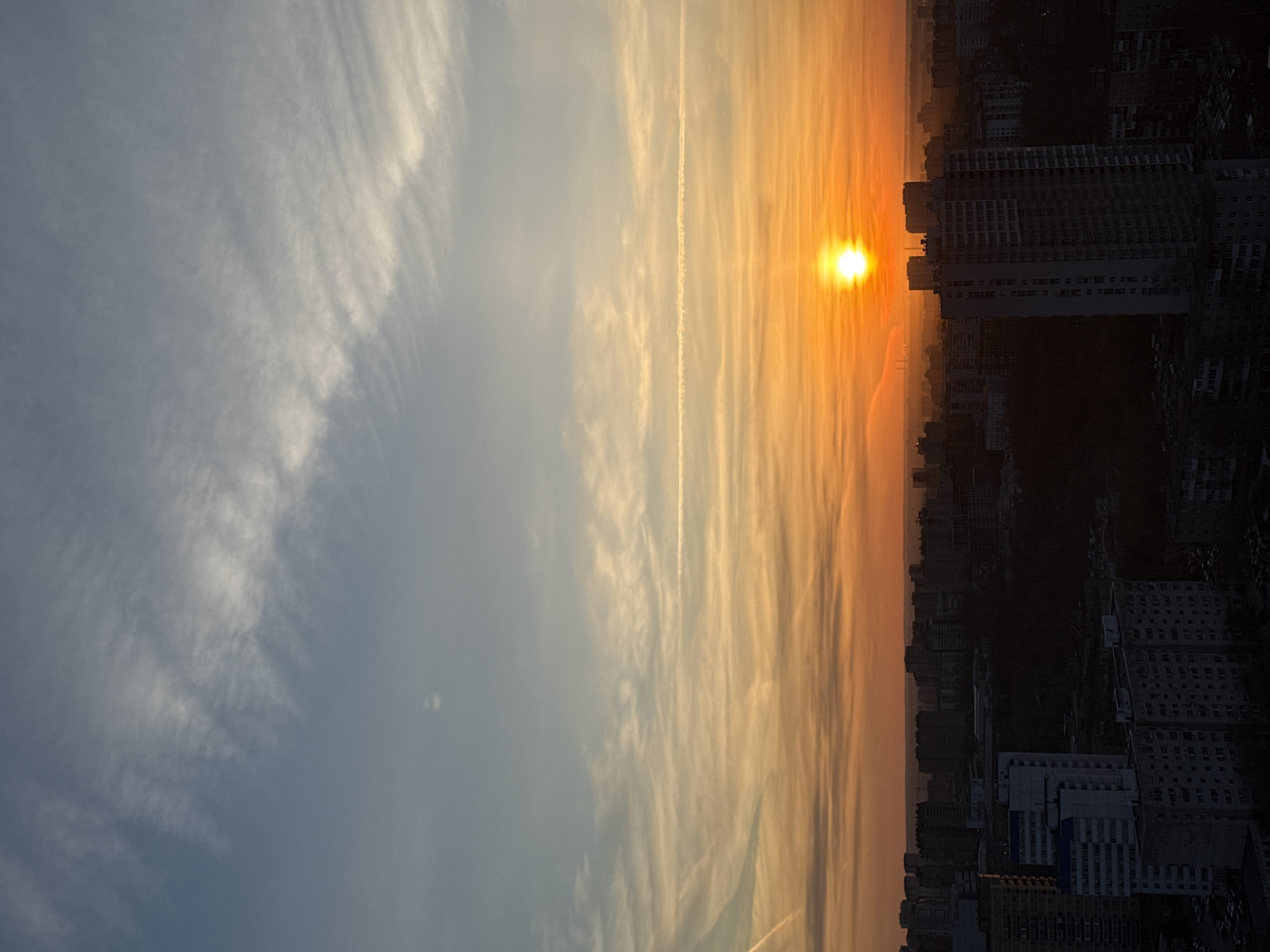 This vertically oriented image captures a vivid sunrise or sunset over the city of Perm, Russia. The scene is dominated by a dramatic sky, transitioning from a soft blue-grey with wispy white clouds in the upper left to a brilliant orange and yellow hue towards the right. A prominent, bright yellow sun, slightly diffused, is positioned in the mid-right portion of the frame, low above the horizon.
A long, thin white contrail stretches horizontally across the sky, cutting through the colorful clouds. Below the vibrant sky, the urban landscape is silhouetted in dark tones against the bright light. Numerous buildings, likely residential or commercial structures, form a dense cityscape across the lower section of the image. One particularly large and dark building stands out on the right, partially obscuring the sun and emphasizing the deep shadows cast by the low-angle light.
The photograph depicts a tranquil moment, likely either dawn or dusk, with atmospheric haze visible near the horizon. There are no people or specific activities depicted. No discernible text is visible within the image.