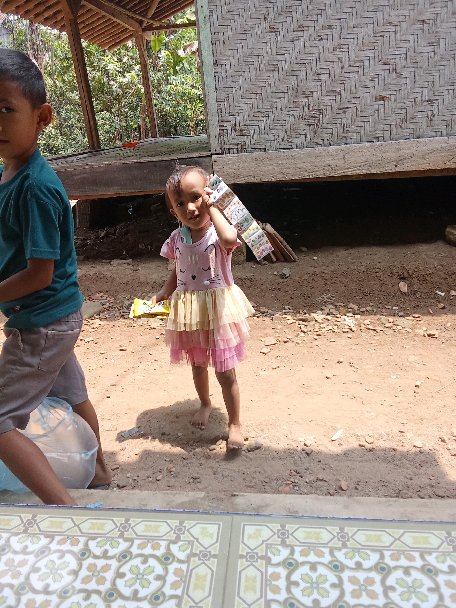 Two children are outdoors near a rustic wooden structure. In the foreground, a young boy is partially visible on the left, wearing a teal shirt and grey shorts, carrying a large translucent plastic bag. To the right of the boy, a young girl stands facing forward, wearing a pink and yellow dress adorned with a cat face design. She is holding a strip of colorful stickers up to her ear, as if on the phone, and has a yellow packet in her left hand. The ground is dirt with scattered pebbles and some debris. The background shows the weathered woven bamboo wall of the structure and lush green foliage. The scene appears to be in a rural setting, potentially the city of Cempaka, Indonesia, given the environmental context and architectural style.