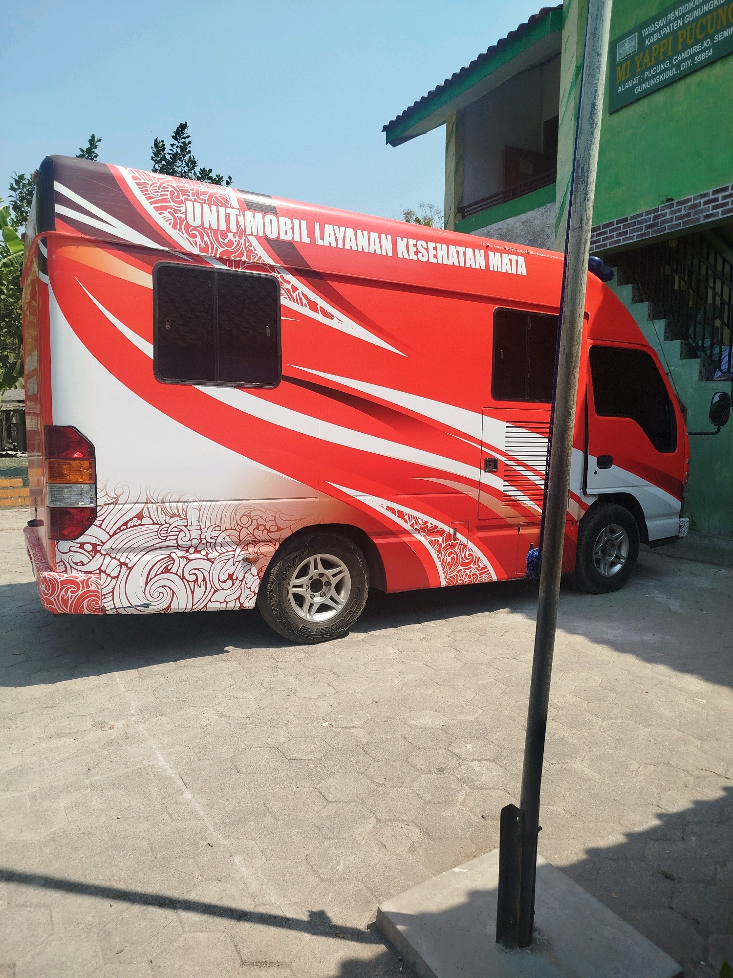 In the city of Semin, Indonesia, a red and white mobile eye health service van is parked on a hexagonal paved surface outdoors during the daytime. The van is adorned with a graphic design that includes swirling red patterns and the text "UNIT MOBIL LAYANAN KESEHATAN MATA" in white. To the right of the van, a green building with a white roof and steps leading up to its entrance is visible. A vertical metal pole stands in front of the building and the van, casting a shadow on the ground. A green sign with white text is affixed to the building, displaying information about the "MT YAPPI PUCUNG" institution, including its address in Semin, Gunungkidul. The sky above is clear and blue.
