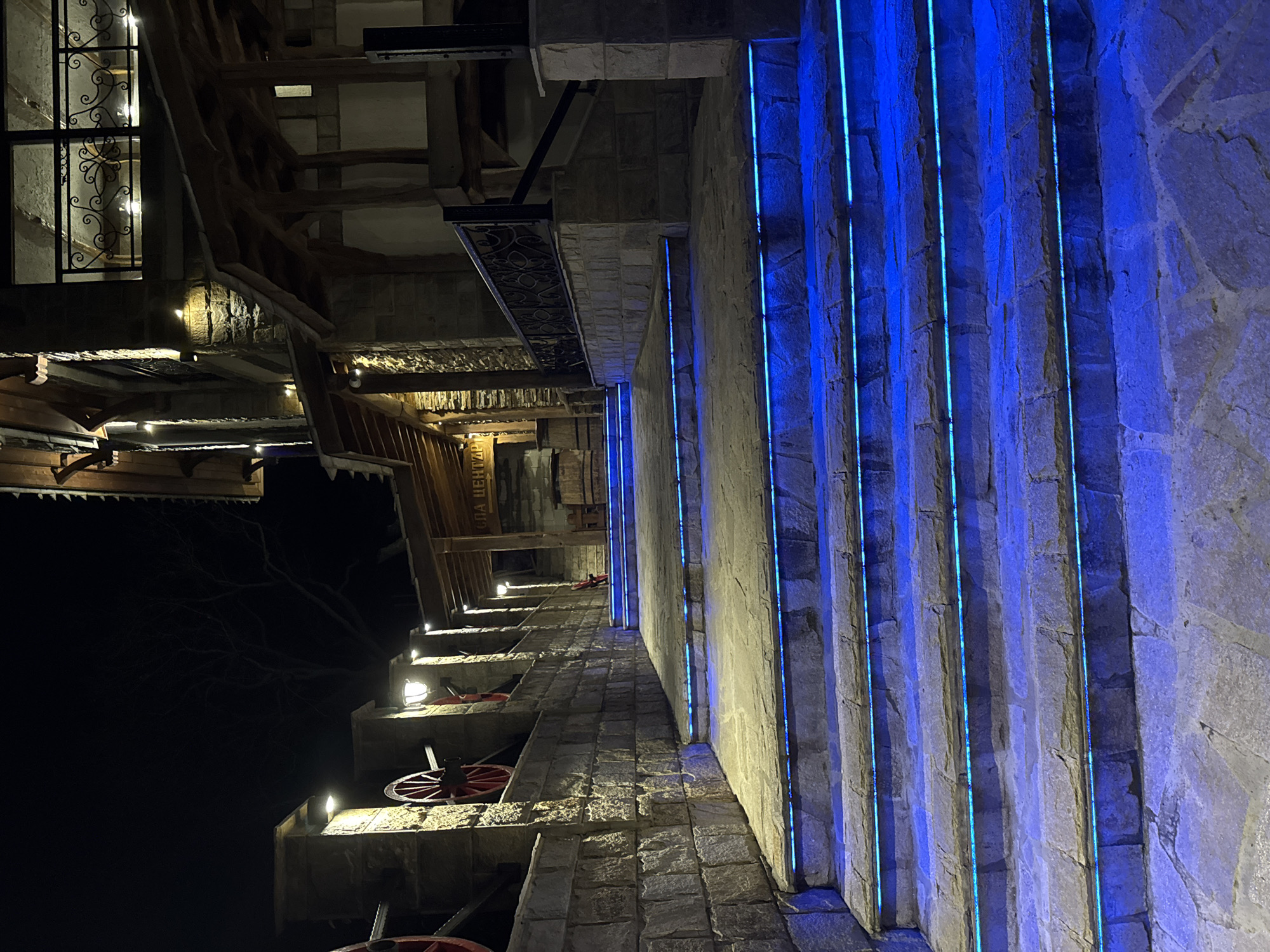 This is an eye-level, outdoor shot in Medovo, Bulgaria, featuring a distinctive stone building illuminated at night. The building appears to be a multi-story structure with a rustic design, incorporating wood and stone elements. On the right, a tall stone wall is adorned with vertical blue neon lights, creating a striking visual contrast against the building's stonework.
On the left, the building has a window with decorative ironwork. Other architectural features include wooden beams, awnings, and what appears to be a staircase or ramp leading up the building. Along this pathway are mounted lanterns or lights illuminating the steps. Red wheels are visible, possibly part of a decorative or functional mechanism attached to the building. The sky in the background is dark, suggesting a nighttime setting.