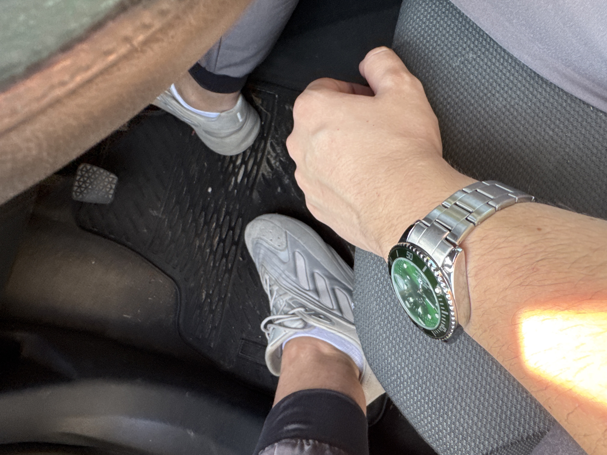 The image shows a close-up view of a person's arm and hand wearing a silver wristwatch with a green face, resting on a textured grey seat. In the background, out of focus, are two pairs of silver athletic shoes and a car's floor mat and pedals. The scene appears to be inside a vehicle. There is no indication of the location being in the city of Madan, Bulgaria.