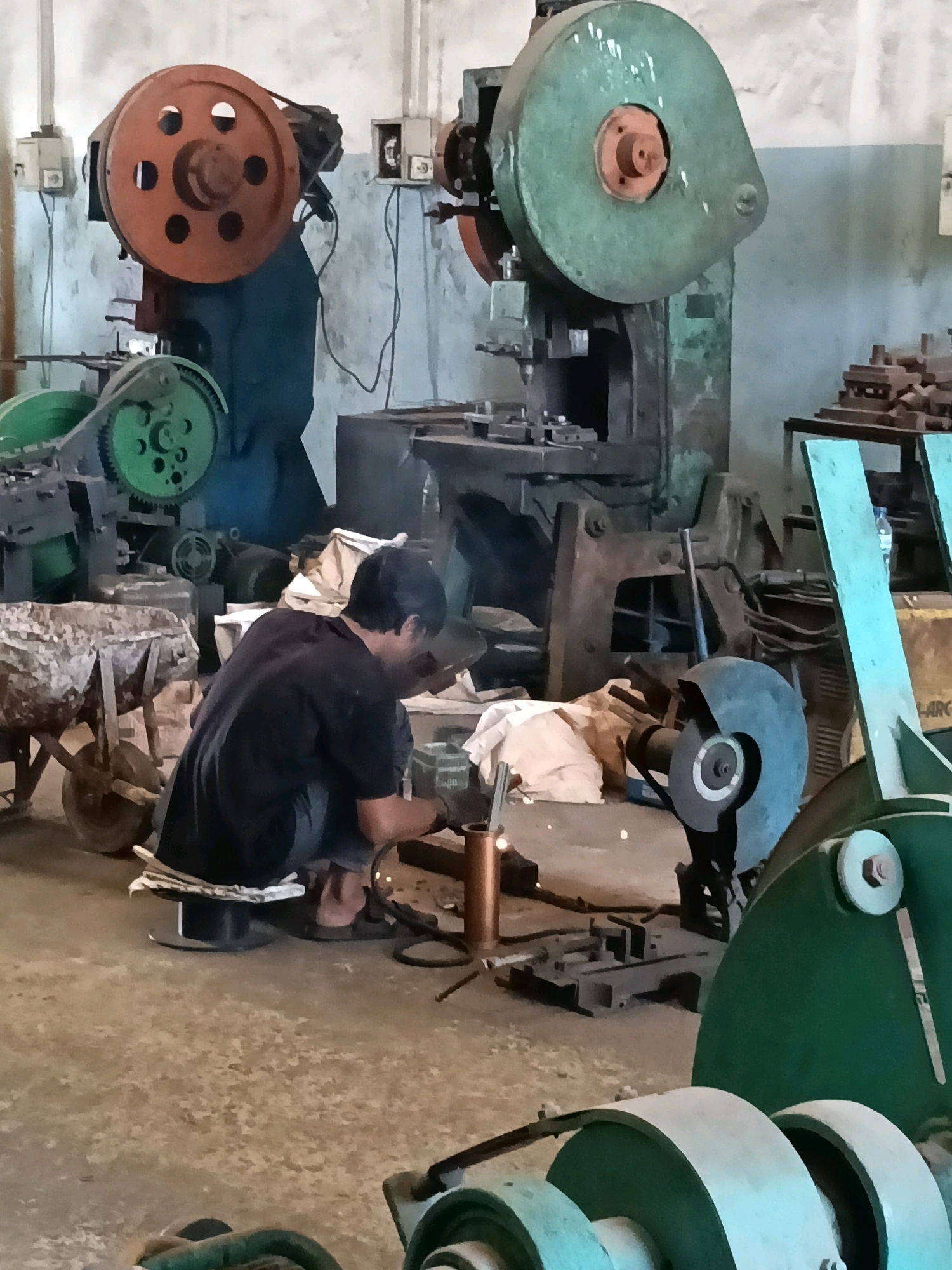 The image depicts an indoor workshop or factory setting, likely in the city of Belimbing, Indonesia, with industrial machinery. In the foreground, a person is crouched on the floor, appearing to be welding or engaged in a similar metalworking activity. The individual is wearing a dark t-shirt and dark pants, with their back mostly to the camera. They are working on a piece of metal, and sparks are visible around the welding area.
Surrounding the individual are various pieces of heavy machinery. To the left, a large, orange-wheeled machine with a dark blue base stands prominently. Further left, a green-geared machine is visible. In the background, a large green industrial press machine with a heavy circular element on top dominates the scene. Other metal components, tools, and materials are scattered throughout the workshop. A light blue-colored piece of equipment is visible on the right side of the frame. The floor is concrete, and the walls appear to be light-colored. The overall impression is one of an active, albeit somewhat cluttered, industrial workspace.
