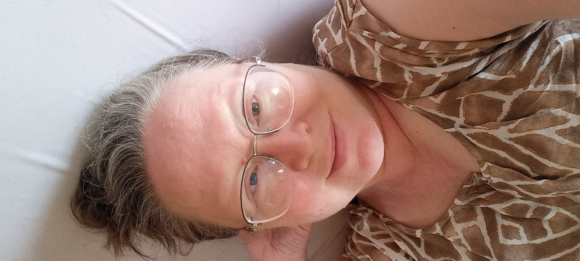 A woman with graying brown hair and glasses is lying down, looking at the camera. She is wearing a brown and white patterned top. Her left hand is behind her head. The background is a light-colored, textured surface, possibly a bed or sofa. The image does not contain any information that would allow for its localization in Izhevsk, Russia, or any other specific city.