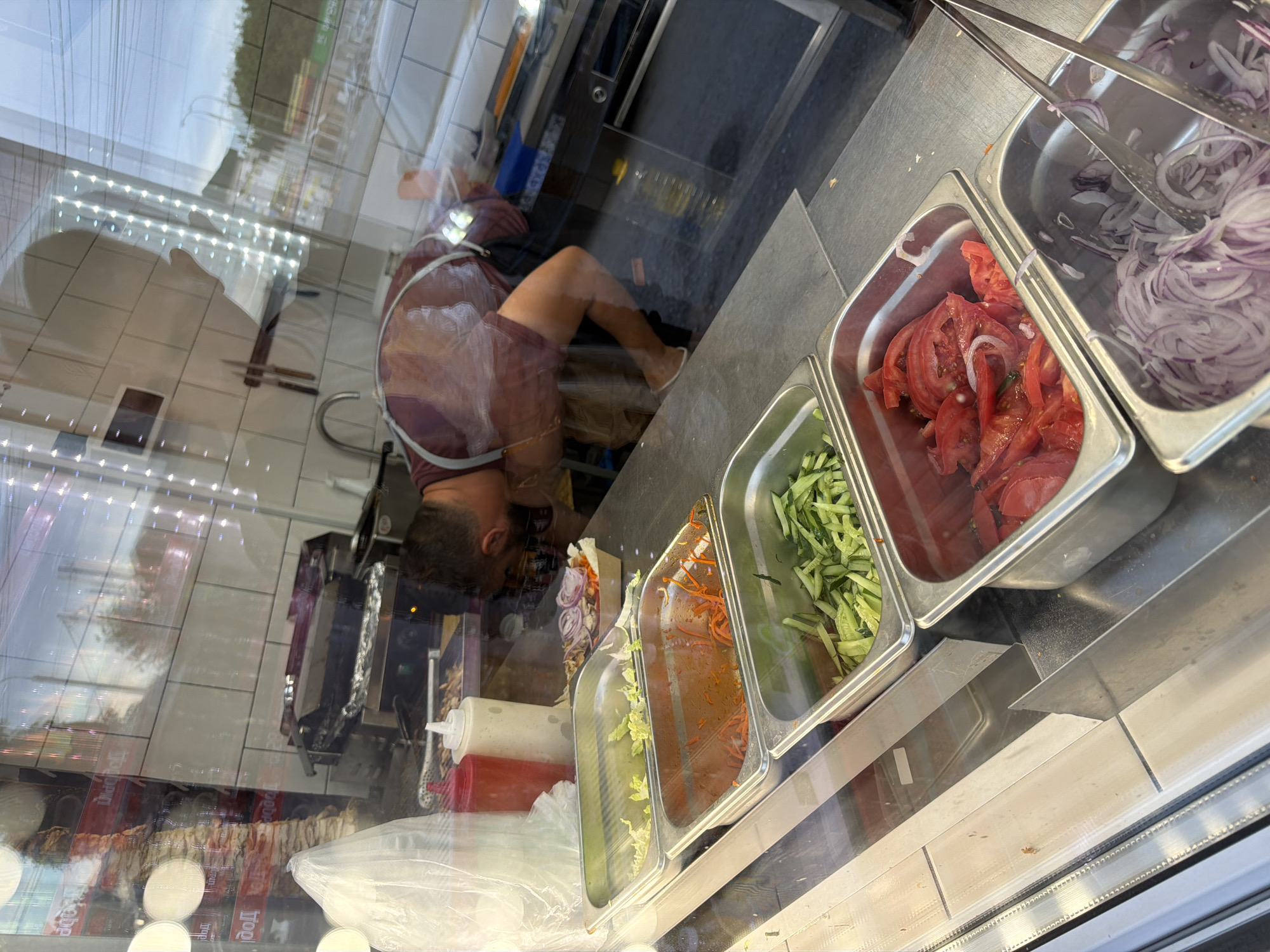 A man with dark hair, wearing an apron over a burgundy shirt, leans over a food preparation area in a street food establishment. He is inspecting the contents of metal trays filled with ingredients. The trays contain shredded lettuce, shredded carrots, sliced cucumbers, sliced tomatoes with onions, and sliced red onions. To the left, there are two squeeze bottles, one red and one white, and a clear plastic bag. The scene appears to be in a city, with reflections of buildings and a street visible in the glass window in front of the food preparation area. This establishment is located in Volzhskiy, Russia.
