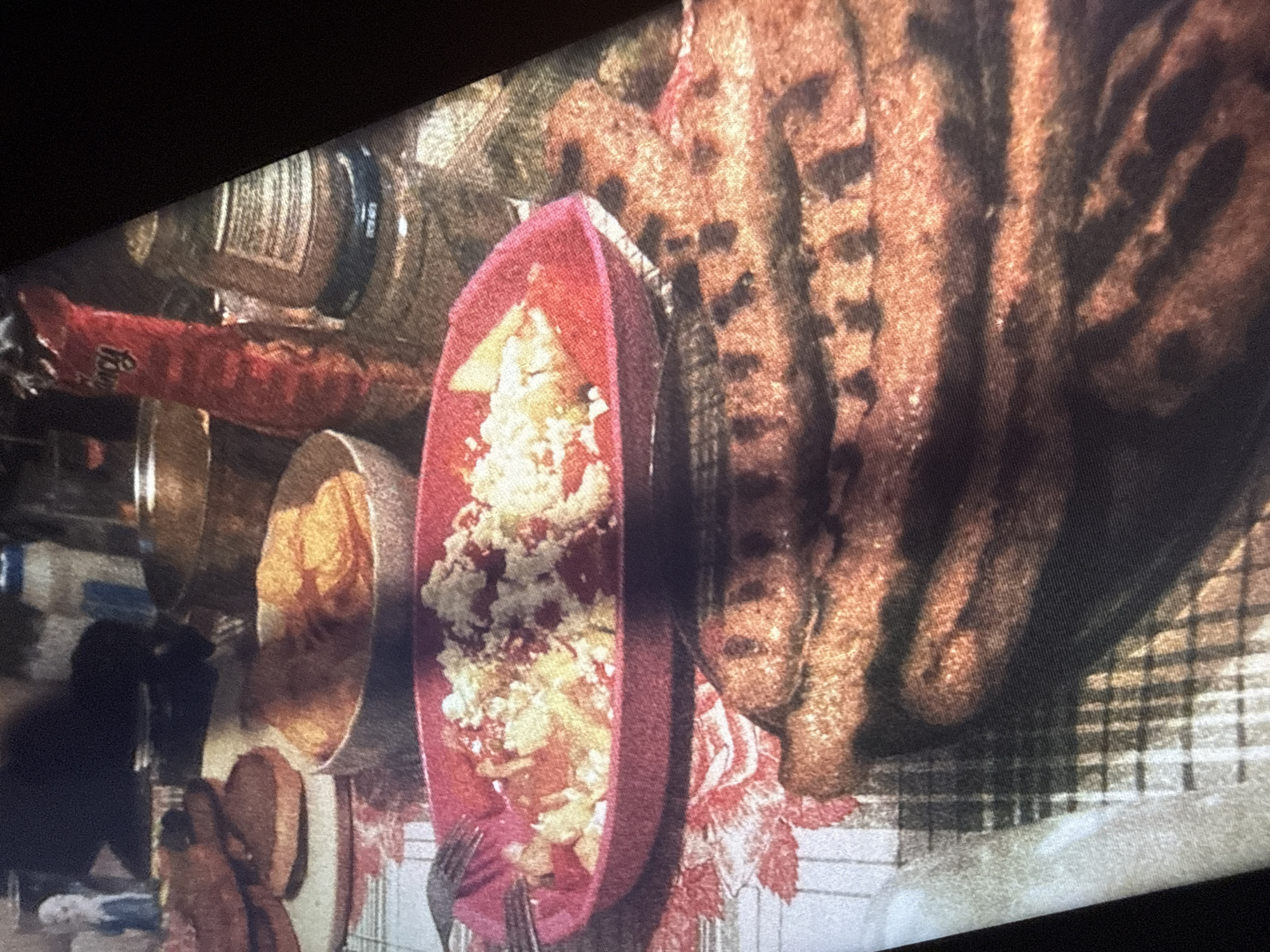 The image displays a close-up of a meal on a table covered with a patterned tablecloth. In the foreground, a stack of grilled sausages, characterized by dark grill marks, is prominently featured. To the left of the sausages, a pink oval dish contains a mixture of what appears to be a salad with cubed vegetables and shredded cheese. A fork rests on the left edge of this dish. In the background, a bowl holds orange potato chips. Behind the bowl, a red squeeze bottle, possibly ketchup, is visible. Further back, a glass jar with a label, likely containing spices or preserves, is situated next to the red bottle. The scene is dimly lit, suggesting an indoor setting, possibly a dining area. There are no discernible landmarks or architectural details that would indicate a specific location in Sofia, Bulgaria. The focus is entirely on the food and table setting.