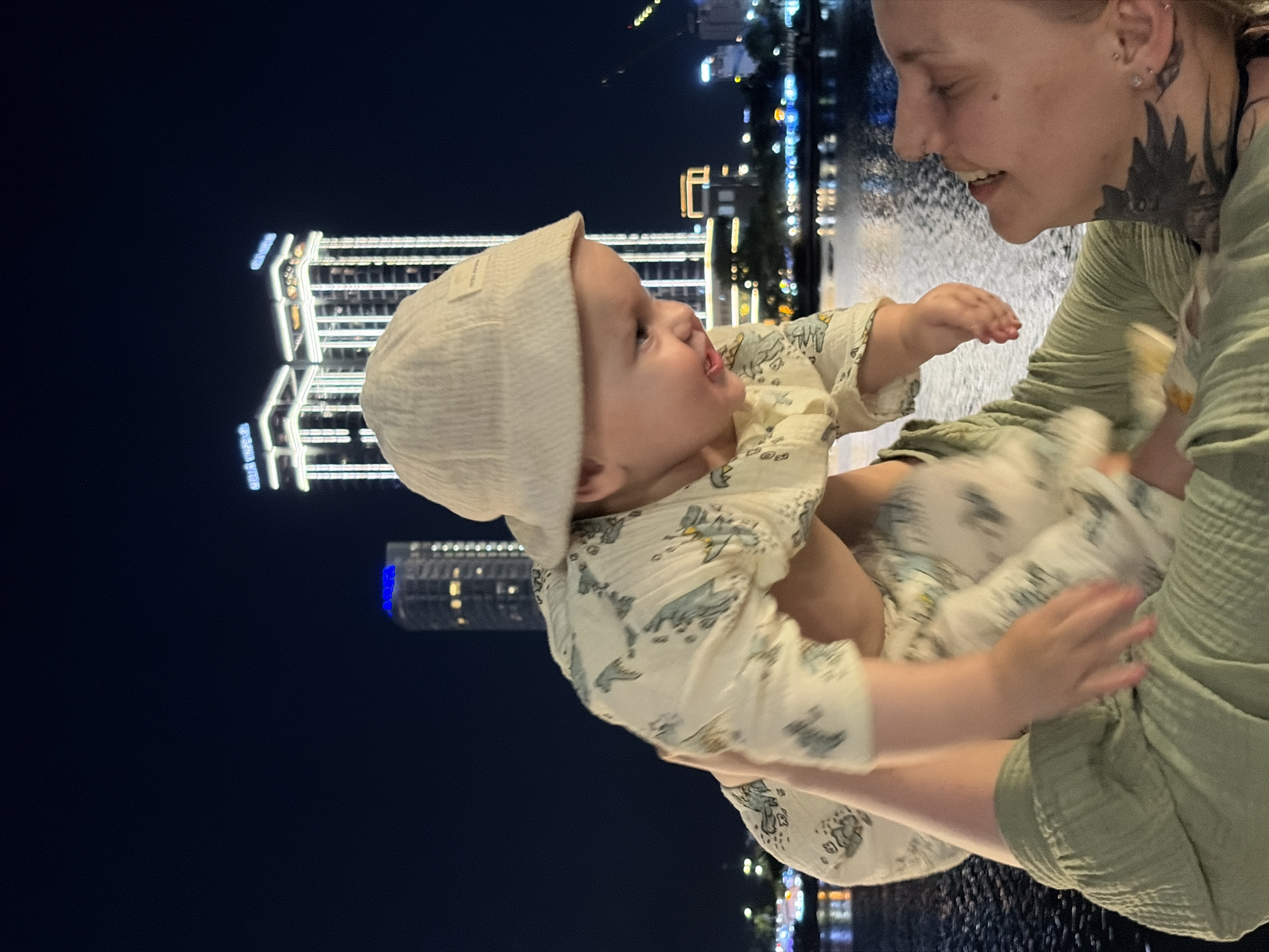 The image captures a heartwarming moment between a woman and a baby at night, set against the illuminated cityscape of Da Nang, Vietnam. The woman, with light skin, ear piercings, and a distinctive dark tattoo on her neck and shoulder, wears a light green, textured top. She is holding the baby, who also has light skin and is dressed in a cream-colored, textured beanie and a light-colored long-sleeved onesie patterned with what appear to be small light blue or grey animals or shapes.
Both subjects are looking at each other with clear expressions of joy and affection. The baby's mouth is open in a wide smile or laugh, with arms raised, seemingly reaching out playfully towards the woman. The woman smiles warmly back, her head tilted slightly, indicating a loving interaction.
The scene is outdoors at night, characterized by a dark sky contrasting sharply with a background of brightly lit modern city buildings. Between the subjects and the buildings, there is a dynamic water feature, likely a fountain or water jets, creating a misty, textured effect that diffuses some of the background lights. Reflections of these urban lights are visible on the wet or watery surface in the foreground.
The time of day is clearly night, as evidenced by the dark sky and abundant artificial illumination from the buildings and water feature. The atmosphere is joyful and intimate, highlighting the bond between the two individuals. On the baby's beanie, a small tag shows faint, indistinct text, possibly a brand name like "TOGAE USA." Reflected text from distant building signage is also visible in the water but is too blurry to decipher.
