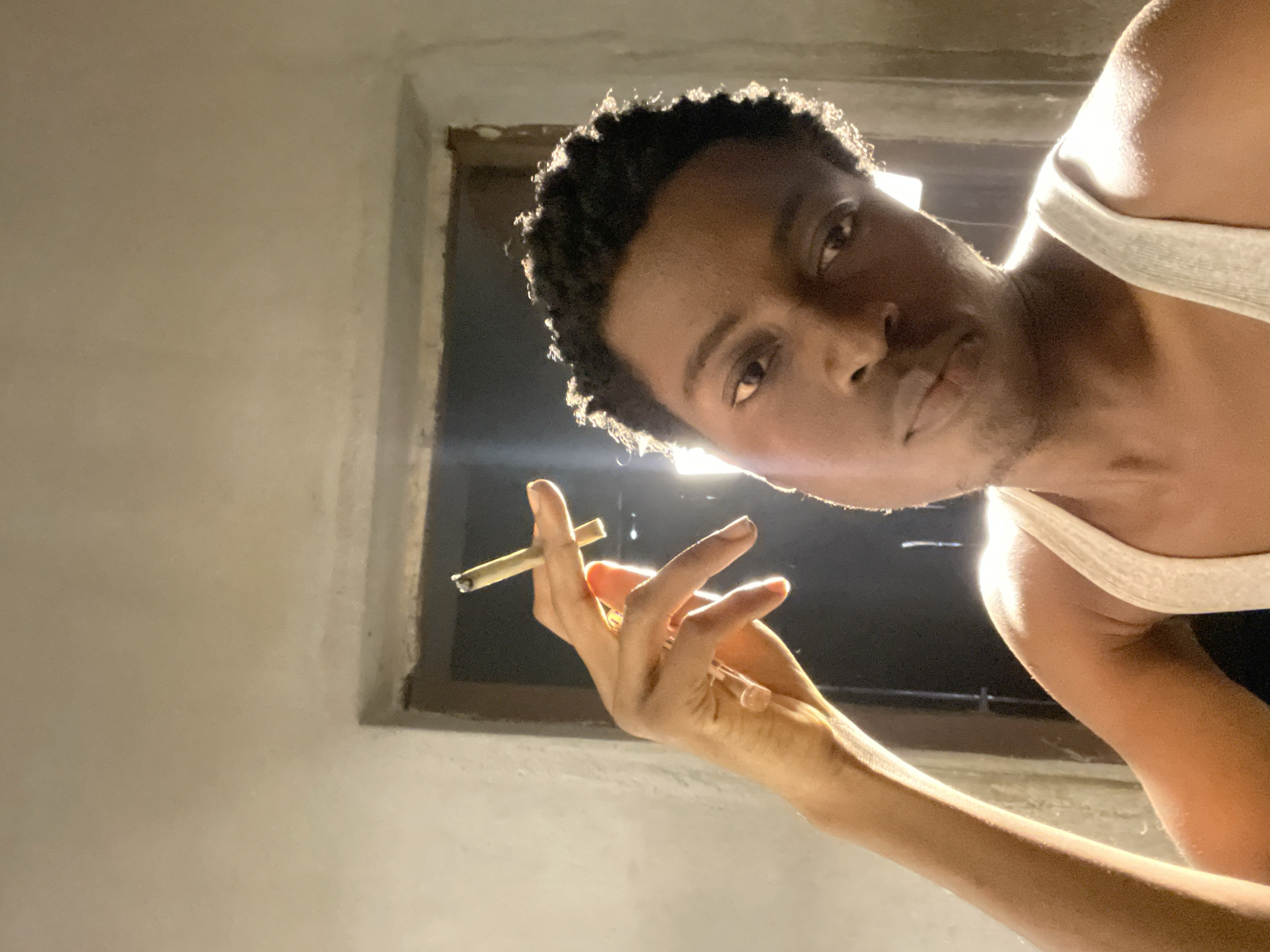 The image is a selfie taken indoors, possibly in Nkpor, Nigeria, as indicated by the context. The subject is a young Black male wearing a white tank top. He is holding a cigarette between the index and middle fingers of his left hand, which is raised near his head. The lighting in the image is dramatic, with a bright light source behind him creating a strong silhouette effect and illuminating the right side of his face and shoulder. The background appears to be a doorway or window with a dark interior visible. The walls are light-colored and textured. The subject has short, curly dark hair and is looking towards the camera with a neutral expression.