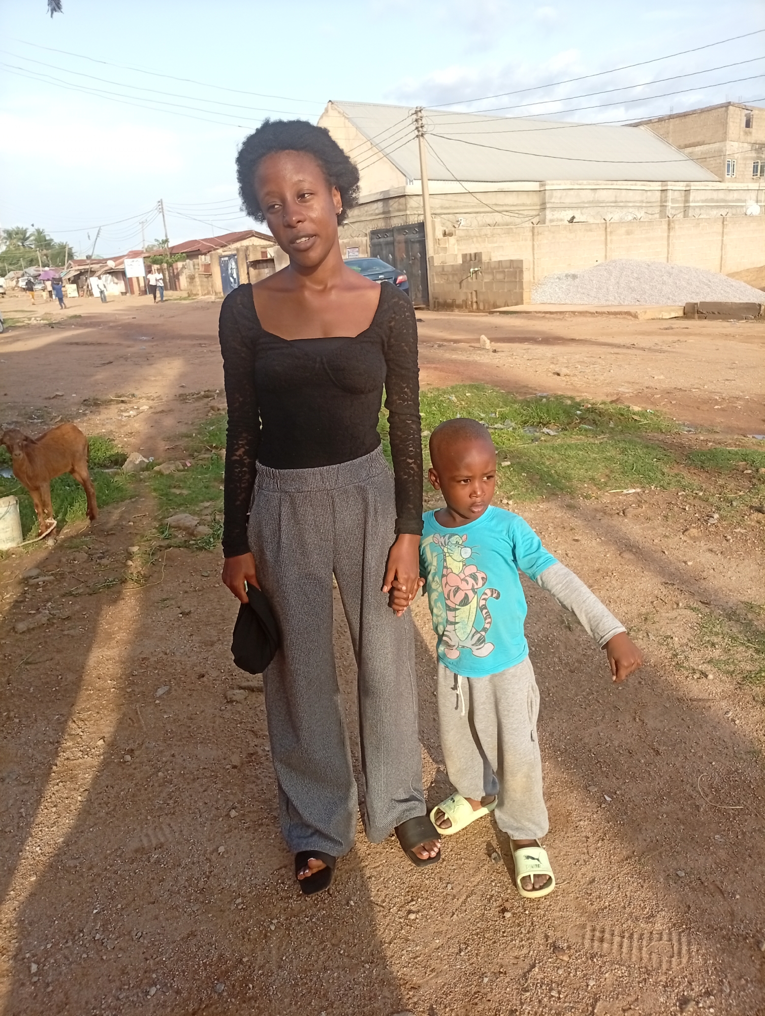 The image depicts a young woman and a child standing side-by-side on a dirt road in Sabon Tasha, Nigeria. The woman is wearing a black long-sleeved top, wide-legged gray pants, and black sandals. She is holding a black hat in one hand and holding hands with the child with the other. The child is wearing a blue, long-sleeved shirt with a tiger cartoon, gray sweatpants, and yellow sandals. The pair are standing on a dirt road. Behind them, there is a goat and a building with a corrugated metal roof and high walls. A pile of gravel is next to the building. In the background are several people walking on the side of the road. Utility poles and power lines are visible in the sky.