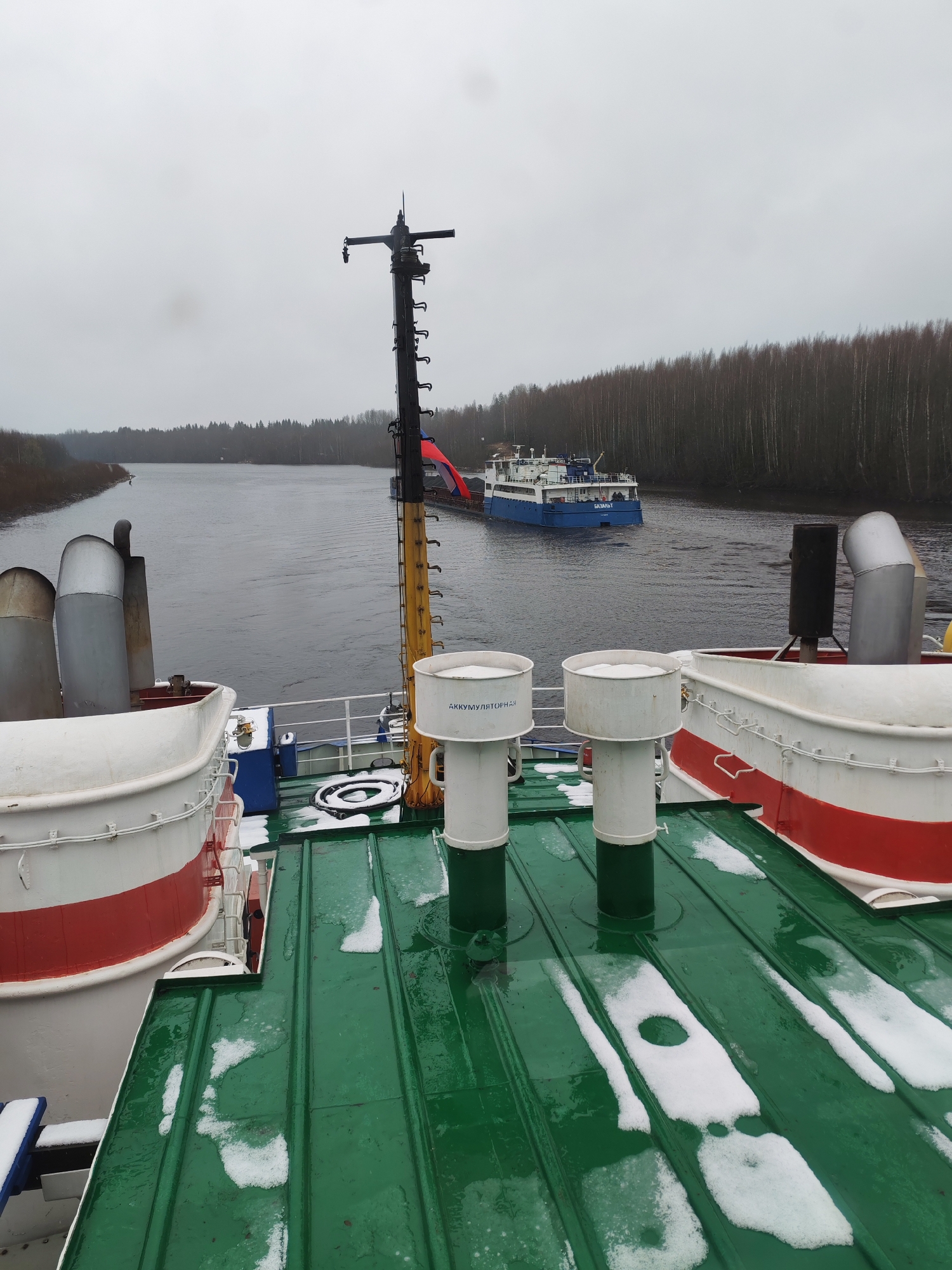 The image shows a view from the deck of a river vessel, likely in the vicinity of the Volokov Most bridge in Russia. The vessel's green deck shows scattered snow. White and red painted structures are visible along the deck's perimeter. A taller, black mast with attached equipment is located mid-frame. In the background, another blue and white vessel is visible on a narrow waterway, bordered by a dense forest on either side under a cloudy, overcast sky.
