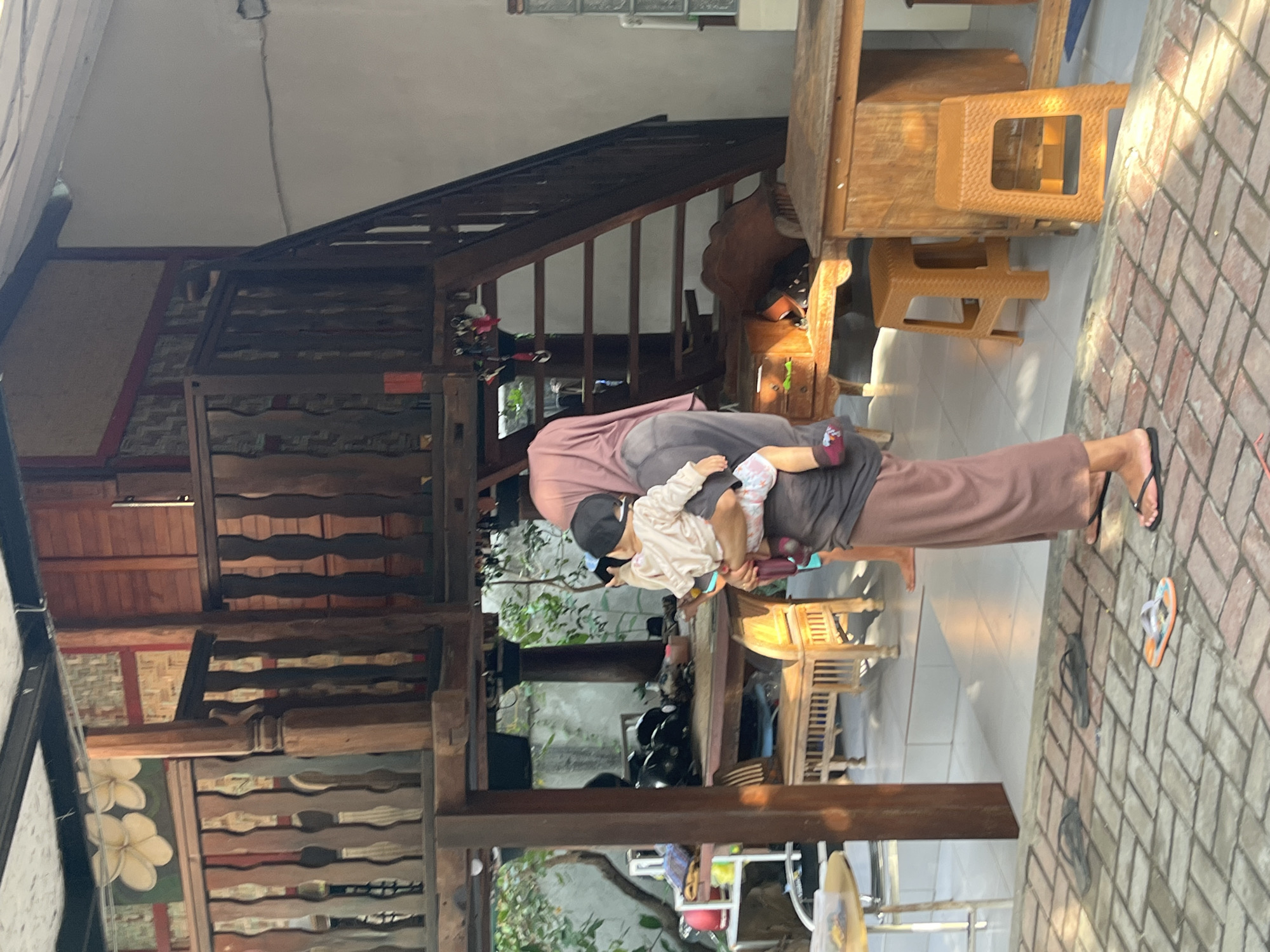 A person, likely a woman wearing a pink headscarf and a long, brown skirt, is holding a young child. The child is wearing a light-colored hooded jacket and a dark baseball cap. They are standing on a brick patio in front of a multi-level wooden structure. The structure features a railing with ornate wooden carvings and a wooden staircase. In the background, there is a table with several black helmets and some greenery. There are also several wooden chairs and stools, as well as a white shelf unit. The scene appears to be outdoors, possibly in a courtyard or a backyard, in Pemenang Timur, Indonesia. The lighting suggests it is daytime, with sunlight casting shadows on the patio.