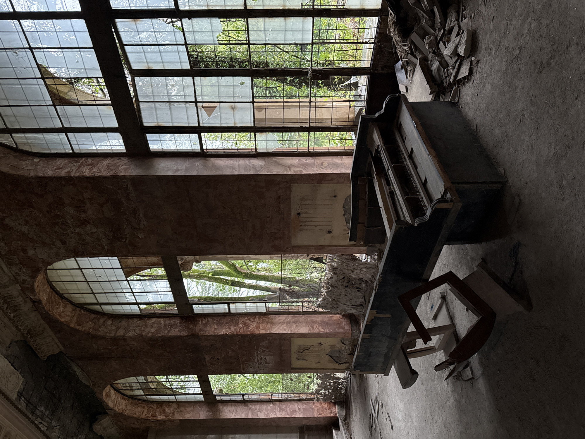 The interior of an abandoned building in Tskaltubo, Georgia is photographed. An old, damaged piano sits on a gray, dirty floor. The walls are brown and peeling, with two tiers of arched windows. Many panes of glass are broken, revealing trees outside. Debris is piled near the piano and by the upper windows. A broken chair is near the piano. The scene is dilapidated and overgrown. No people are visible.