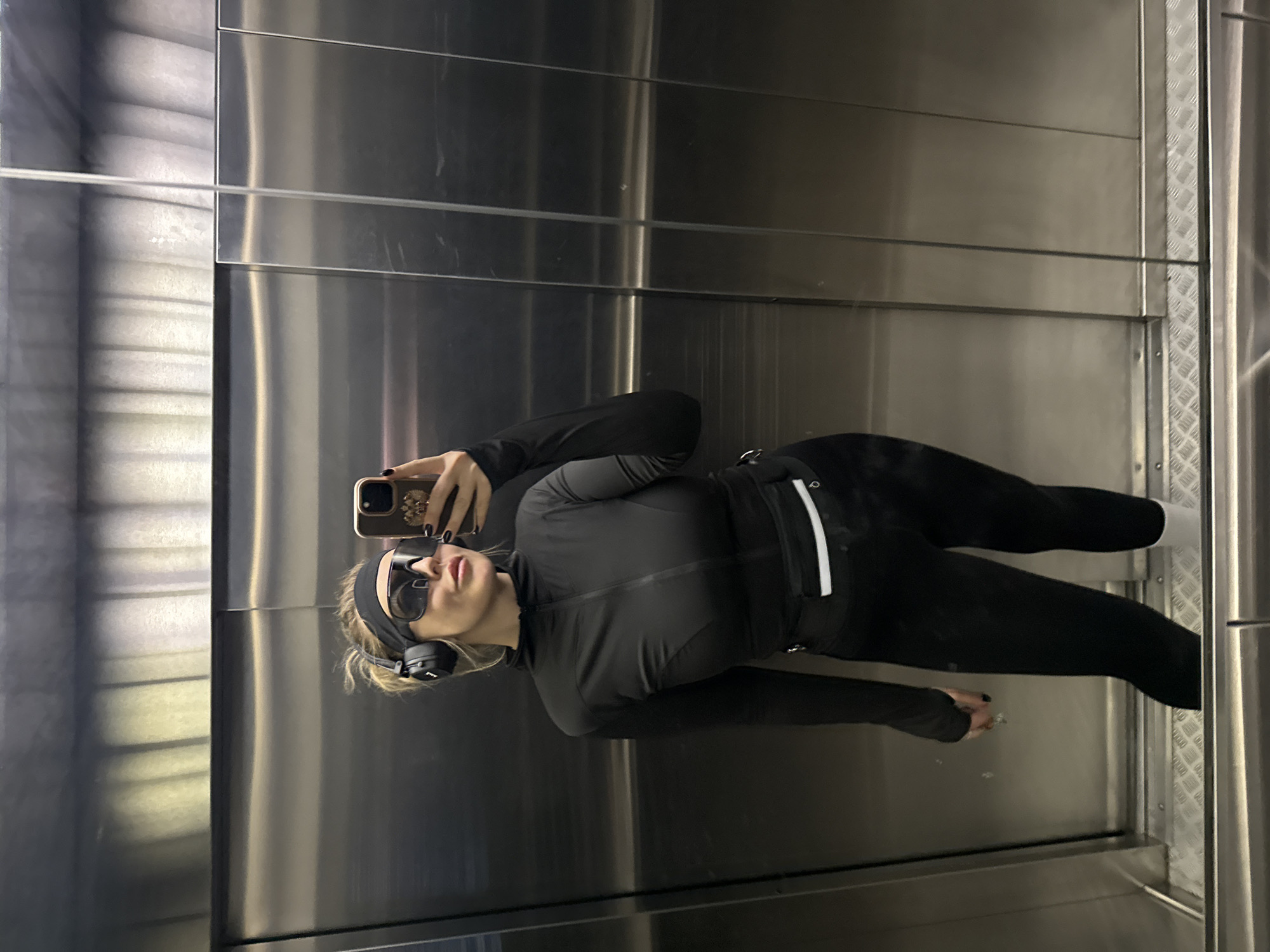 A woman is standing in an elevator, taking a selfie in the mirrored walls. She is wearing a black tracksuit, headphones, sunglasses, and a headband. She holds a smartphone in her right hand. The elevator has silver metallic walls and a textured floor. The location is possibly in Krasnoyarsk, Russia, but this is not verifiable from the image.