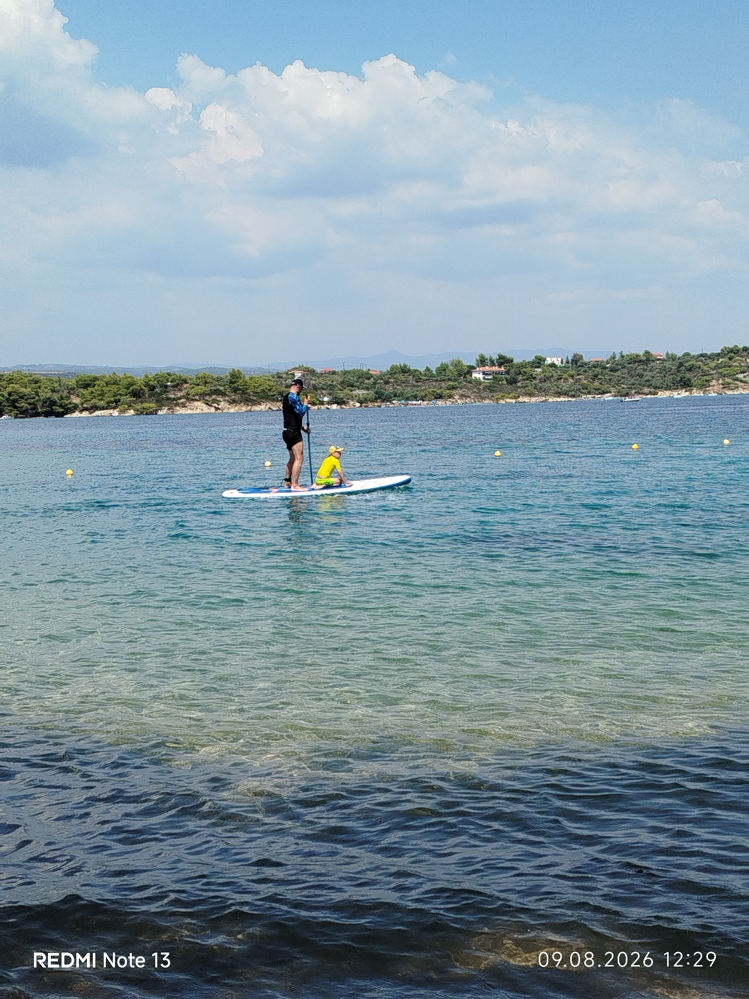 A person on a paddleboard with a child on the same board is in the water at Sithonia Municipal Unit, Greece. The person, wearing a dark rash guard and black shorts, stands on the board, appearing to paddle. The child, wearing a yellow top and shorts, sits on the front of the board. They are on a clear blue body of water with a sandy bottom visible in the foreground. In the background, a tree-covered coastline with buildings is visible under a bright sky with scattered clouds. Three yellow buoys are in the water to the left and right of the paddleboard. The bottom of the image contains a watermark indicating "REDMI Note 13" and a date and time stamp "09.08.2026 12:29".