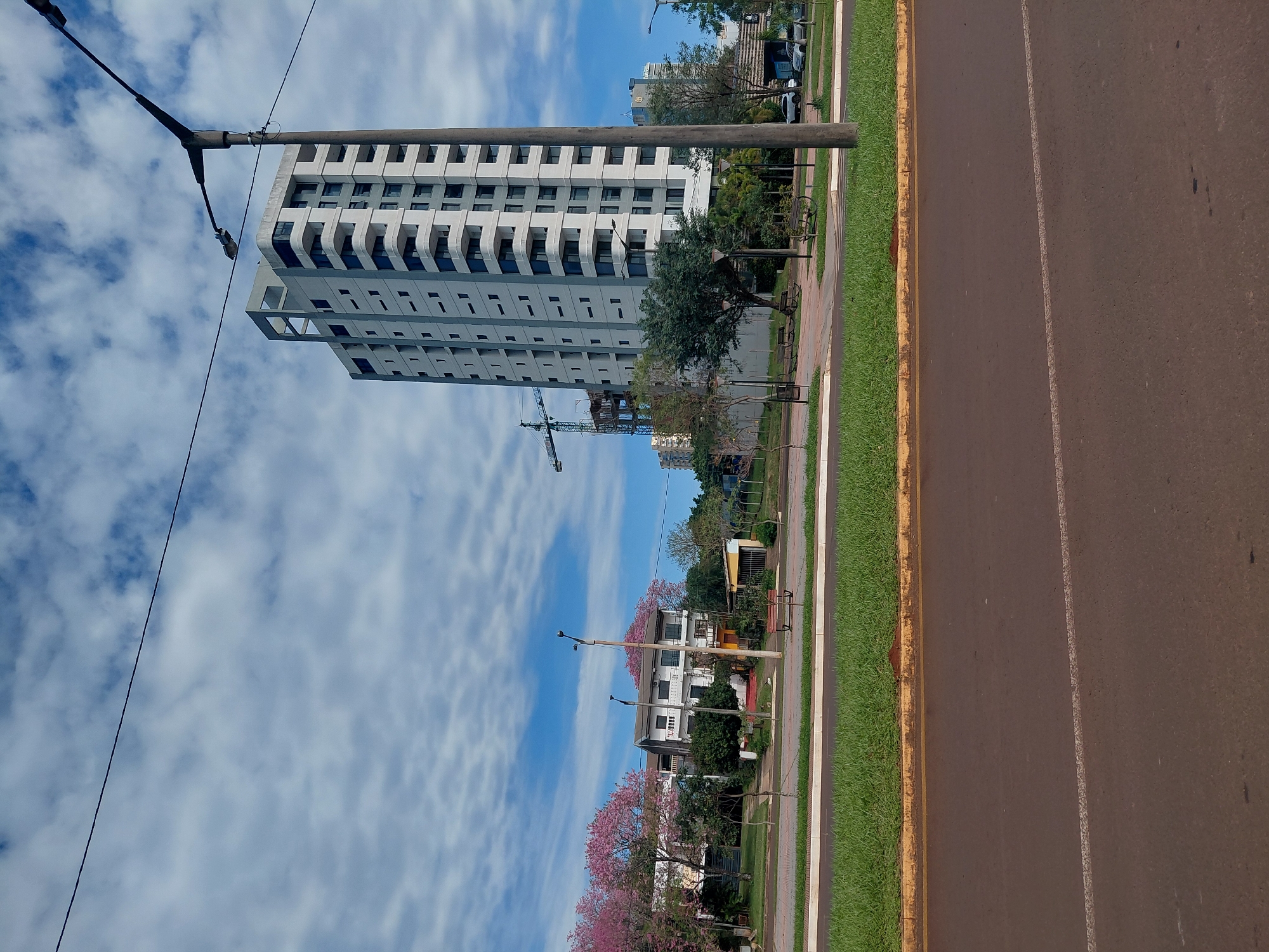 The image captures a street view in Encarnación, Paraguay, under a partly cloudy blue sky. In the foreground, a dark asphalt road with a white lane marker runs horizontally. A grassy median with a yellow curb separates the road from a sidewalk area.
Further back, a row of manicured green grass borders a paved walkway with benches. Beyond the walkway, several trees, some with vibrant pink blossoms, line the path. A low-rise white building with multiple balconies stands behind the trees.
Dominating the mid-ground and background is a tall, modern, white apartment building with numerous balconies and dark-tinted windows. A construction crane is visible to the left of this building, suggesting ongoing development. Another, shorter building is visible behind it. A tall, concrete lamppost with two lights stands prominently in the right mid-ground, with a thin wire running across the sky. Cars are parked and driving in the distance on the right side of the frame. The overall impression is of a well-maintained urban environment with a mix of residential and commercial structures.