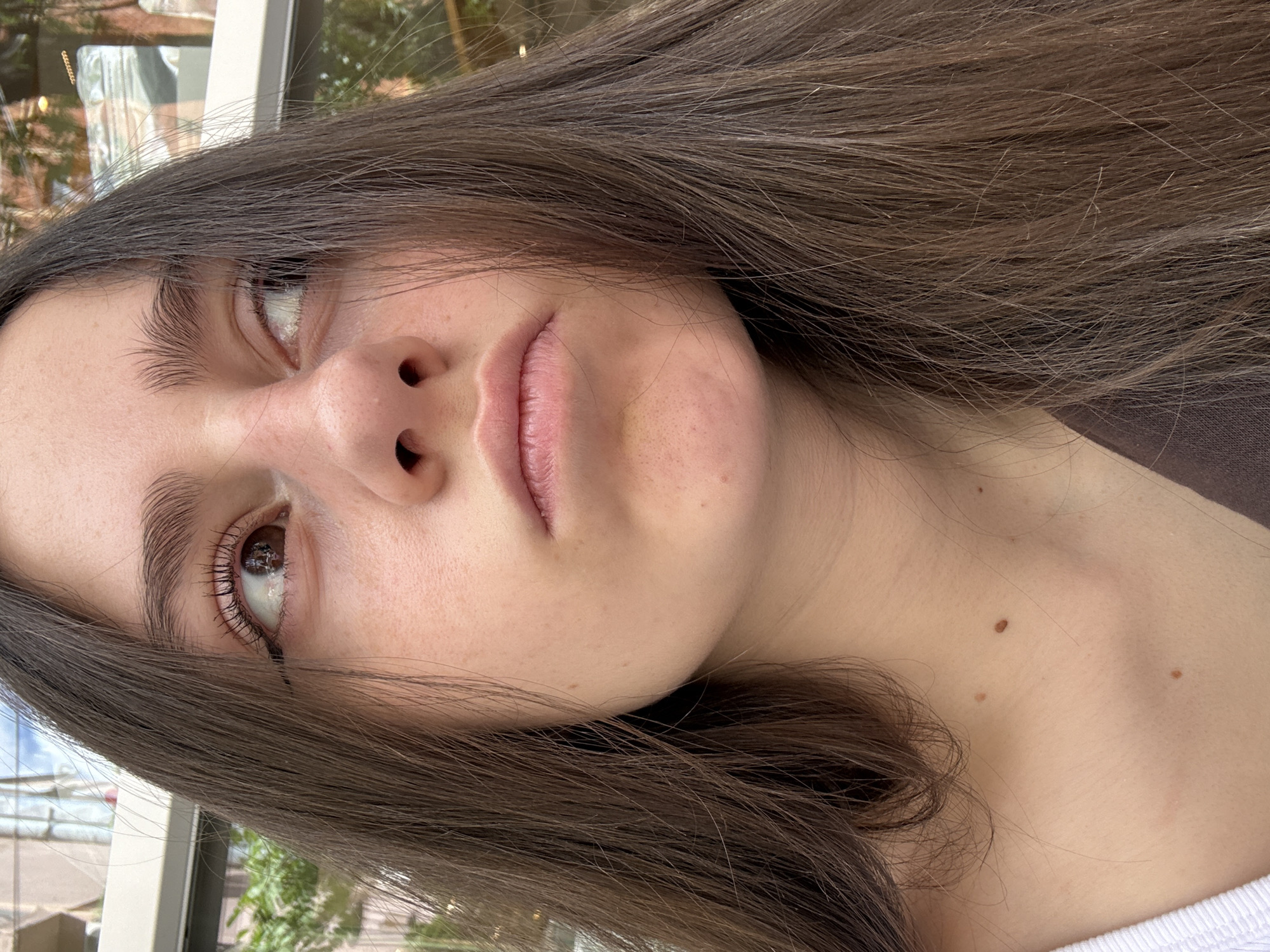 A close-up portrait of a young woman with brown hair, fair skin, and brown eyes. She is looking upwards to the left. Her face is partially illuminated. She has full lips, and her eyebrows are slightly arched. A mole is visible on her neck and chest. The background appears to be a blurry outdoor scene, possibly with greenery and windows, suggesting an urban setting. There is no verifiable information in the image to confirm the location is Yerevan, Armenia. The image provides no direct forensic evidence.