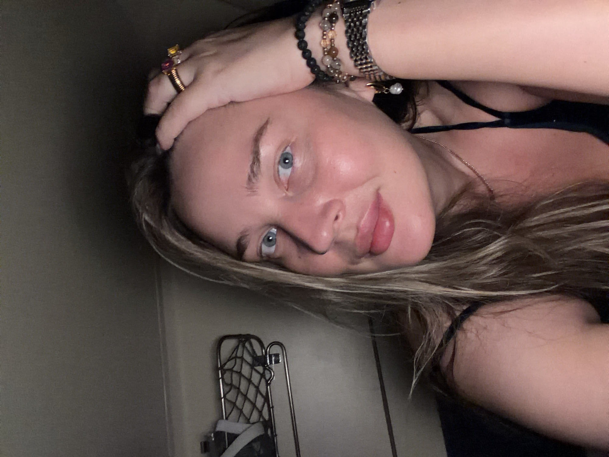 A fair-skinned person with blondish-brown hair, wearing jewelry including rings and bracelets, is positioned close to the camera, looking at the viewer. The background includes a wall, a metal folding chair or drying rack, and a hint of dark clothing. The image's orientation is tilted. The location, Krasnyi Yar, Russia, cannot be confirmed from the image content.