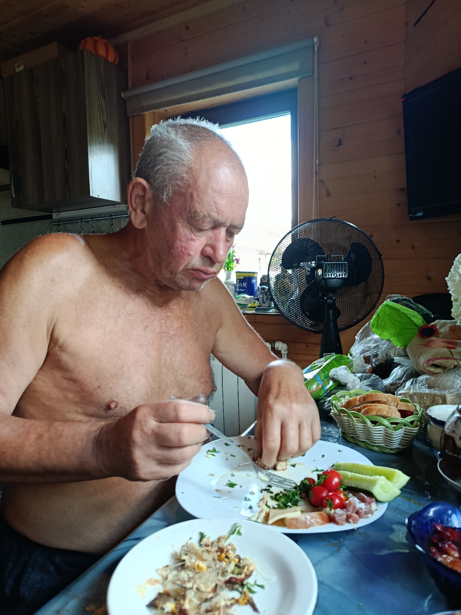 An elderly, shirtless Caucasian man with gray hair is seated at a table indoors, eating. He is using a fork and knife to eat from a white plate that contains food. The table is covered with a blue tablecloth. On the table, there are several plates of food, a basket of bread slices, a bowl of berries, and a black electric fan. The room has wooden walls and a window with a view of what appears to be an outdoor area. The man has visible skin redness on his face and chest. A dark television is mounted on the wall behind him. The overall impression is a casual, domestic meal setting.