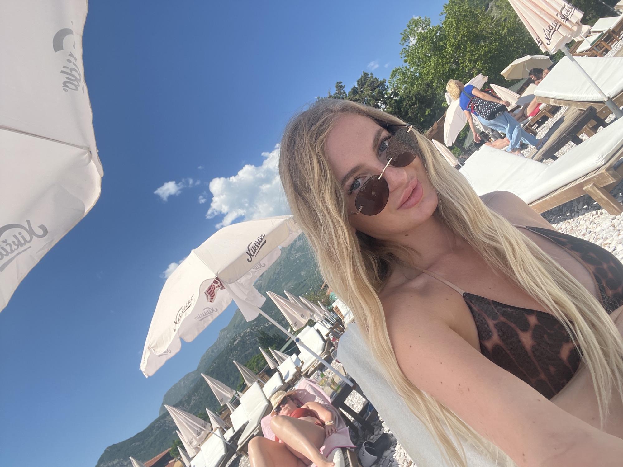 A selfie photo shows a blonde woman with sunglasses on a beach with multiple beach chairs and umbrellas. In the background, mountains are visible. The woman is wearing a brown bikini top. Another person is lying on a beach chair nearby, wearing a hat and a red swimsuit. Other beach patrons are visible in the background. The location appears to be a beach in or near Budva, Montenegro.