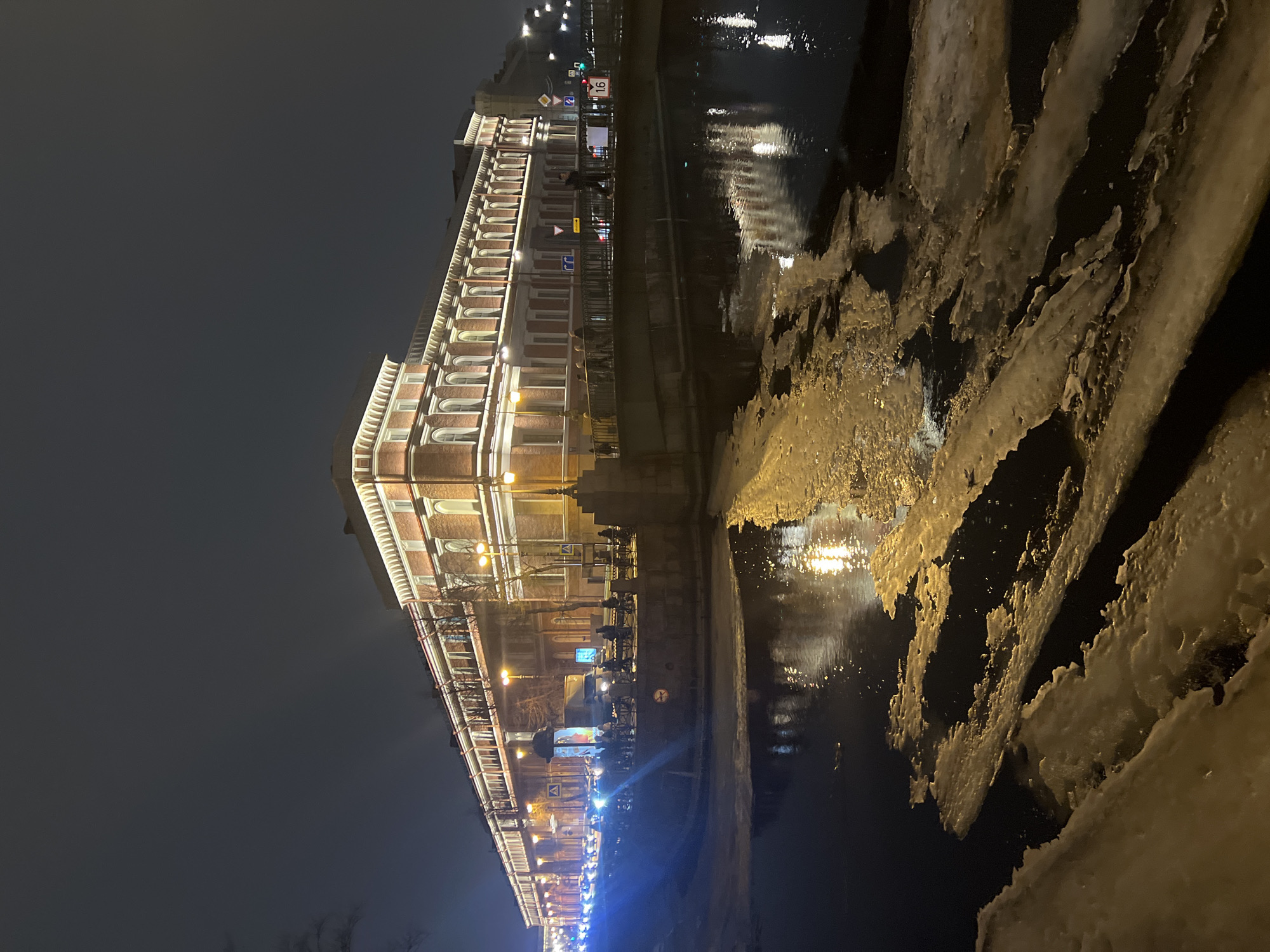 The image presents a nighttime scene along the Kryukov Canal in Saint Petersburg, Russia, depicting the historic buildings of New Holland Island or adjacent structures illuminated against a dark sky. In the foreground, the Kryukov Canal's dark water is partially covered by numerous irregular, yellowish-white ice floes, reflecting the artificial light from the surrounding environment. A dark, metal-railed bridge, likely the Matveev Bridge, crosses the canal in the midground, featuring a red and white sign marked "16" on its structure to the right. Approximately 5-10 indistinct pedestrians are visible walking on the bridge and the adjacent embankment sidewalk. Traffic, including several vehicles with bright blue and white headlights and a light-colored van, proceeds along the street bordering the canal. This street is illuminated by streetlights casting a yellow-orange glow, and an ornate lamppost stands near the bridge. The prominent architectural subject is a long, multi-story building with a reddish-brown brick facade and extensive white architectural detailing, including arched windows and cornices. This building is brightly illuminated by upward-facing lights, emphasizing its features and reflecting onto the canal. Bare tree branches are discernible against the building and along the embankment. The sky above is dark and overcast.