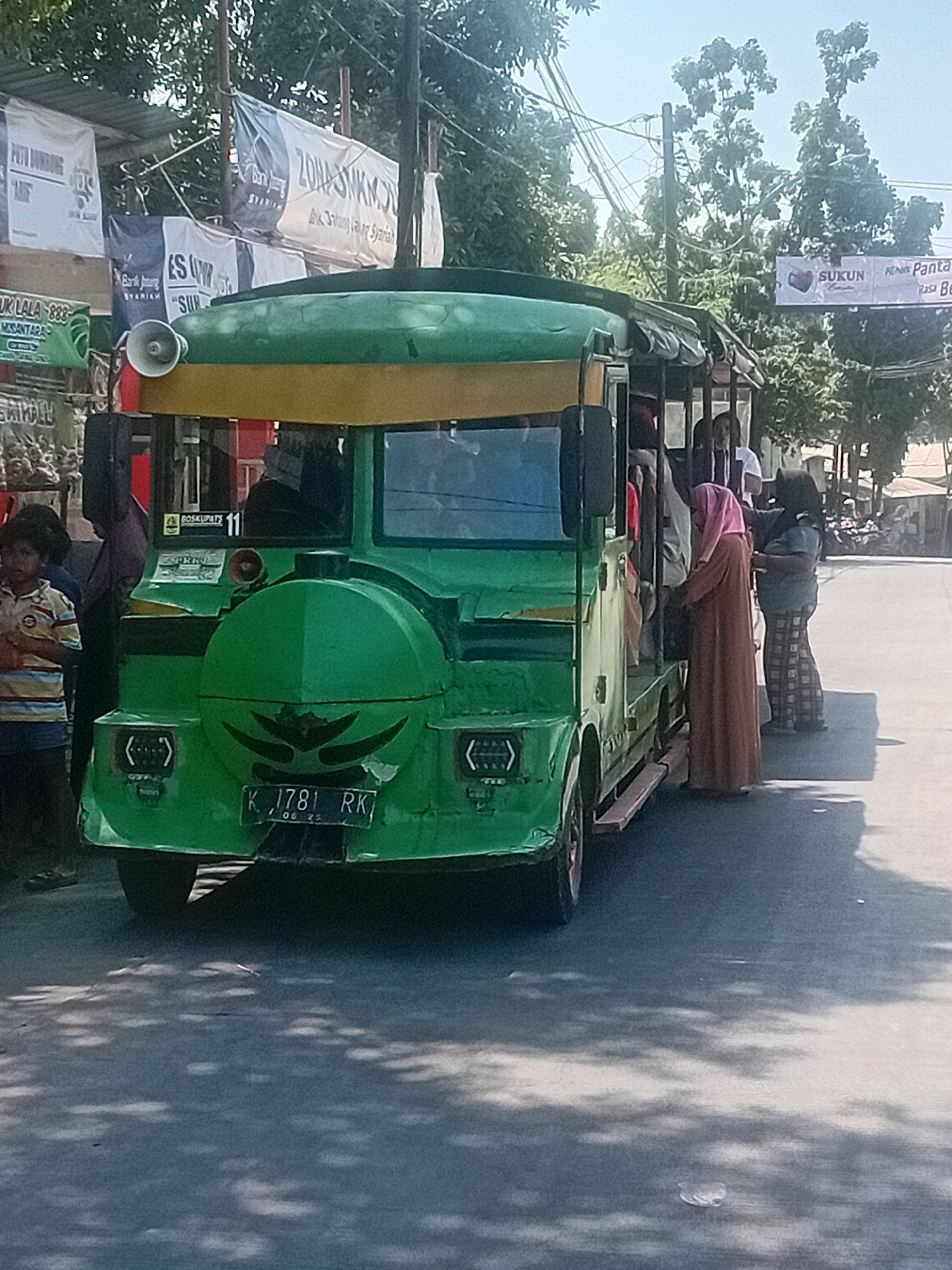 A green and yellow open-sided passenger vehicle, resembling a modified bus or truck, is parked on a paved street in Gribig, Indonesia. The vehicle has a spare tire mounted on the rear, which is green and features a black tribal design. The license plate reads "K. 1781 RK". Several people are visible around and inside the vehicle. A young boy in a striped shirt stands to the left, facing the vehicle. Inside, a woman in a long brown garment and headscarf is stepping into the vehicle, while another person is partially visible behind her. To the right, a woman in a dark shirt and plaid pants stands near the entrance. Banners are hanging in the background, one of which has text that appears to be in Indonesian and mentions "SUKUN". The environment suggests a sunny day with shadows cast on the road from overhead foliage.