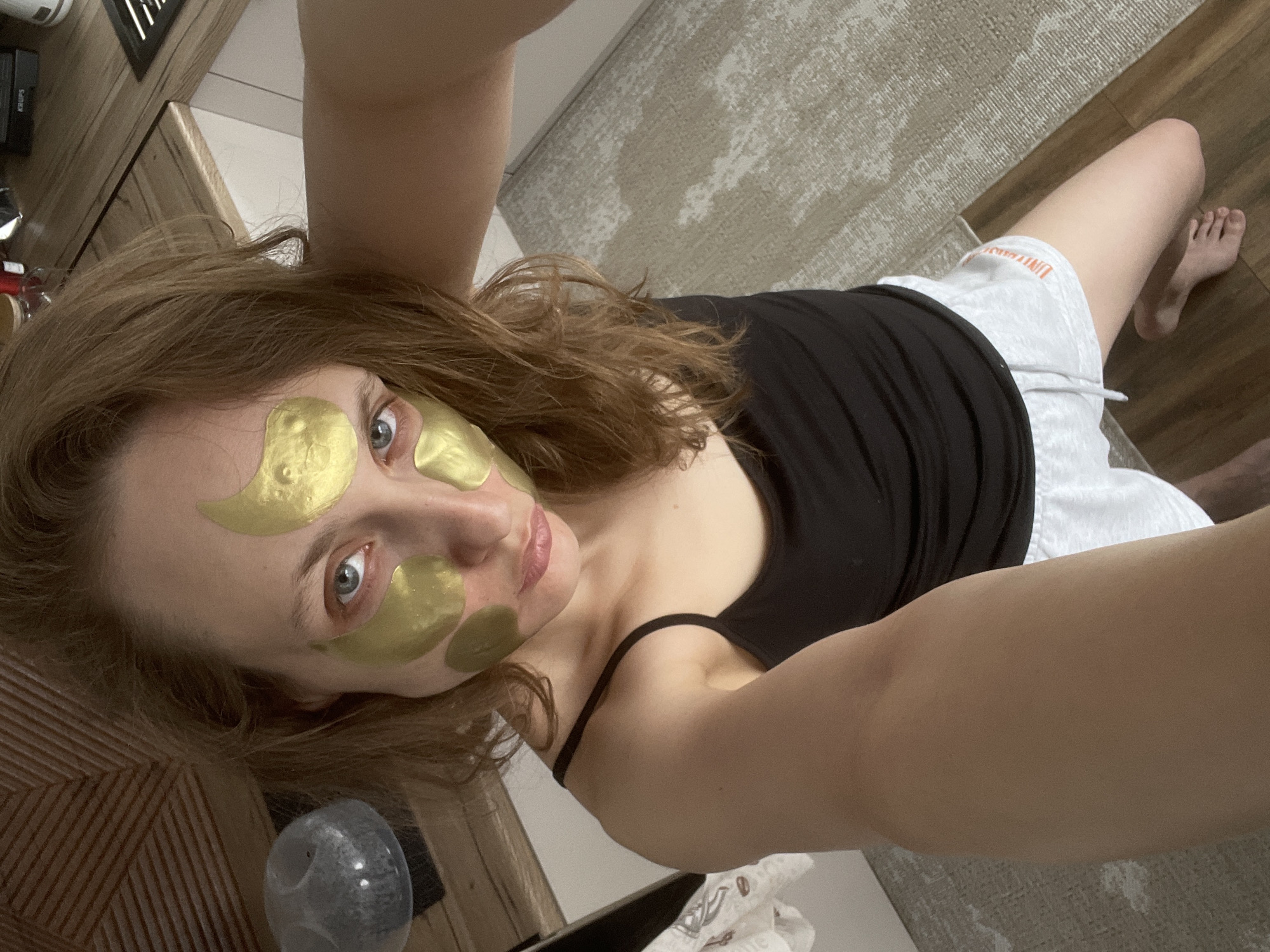 A young woman is captured in a selfie, lying down and looking directly at the camera with a calm expression. She has light brown hair and striking blue eyes. Her face is adorned with several gold hydrogel patches – two under each eye and two on her forehead – indicating a skincare routine. She is wearing a black tank top or camisole and light grey shorts. Visible on the left leg of her shorts is orange text that appears to read "UNIVERSITY." Her lips have a subtle gloss.
The setting is indoors, suggesting a home environment in Podshivalovo, Russia. Her body rests partially on a light-colored, patterned rug, which transitions into a darker wooden floor. Her bare feet are visible towards the right edge of the frame, resting on the wooden floor. To the upper left, behind her head, a wooden surface (possibly furniture) is visible, with blurred objects on top, including what appears to be a black appliance with the brand name "KRUPS" discernible. Near her head, on the wooden surface, there's a translucent, greyish spherical object with a textured surface, possibly a decorative item or a small humidifier. The lighting is soft and even, typical of indoor illumination, and does not provide cues for the time of day. The overall scene conveys a moment of relaxation and self-care.