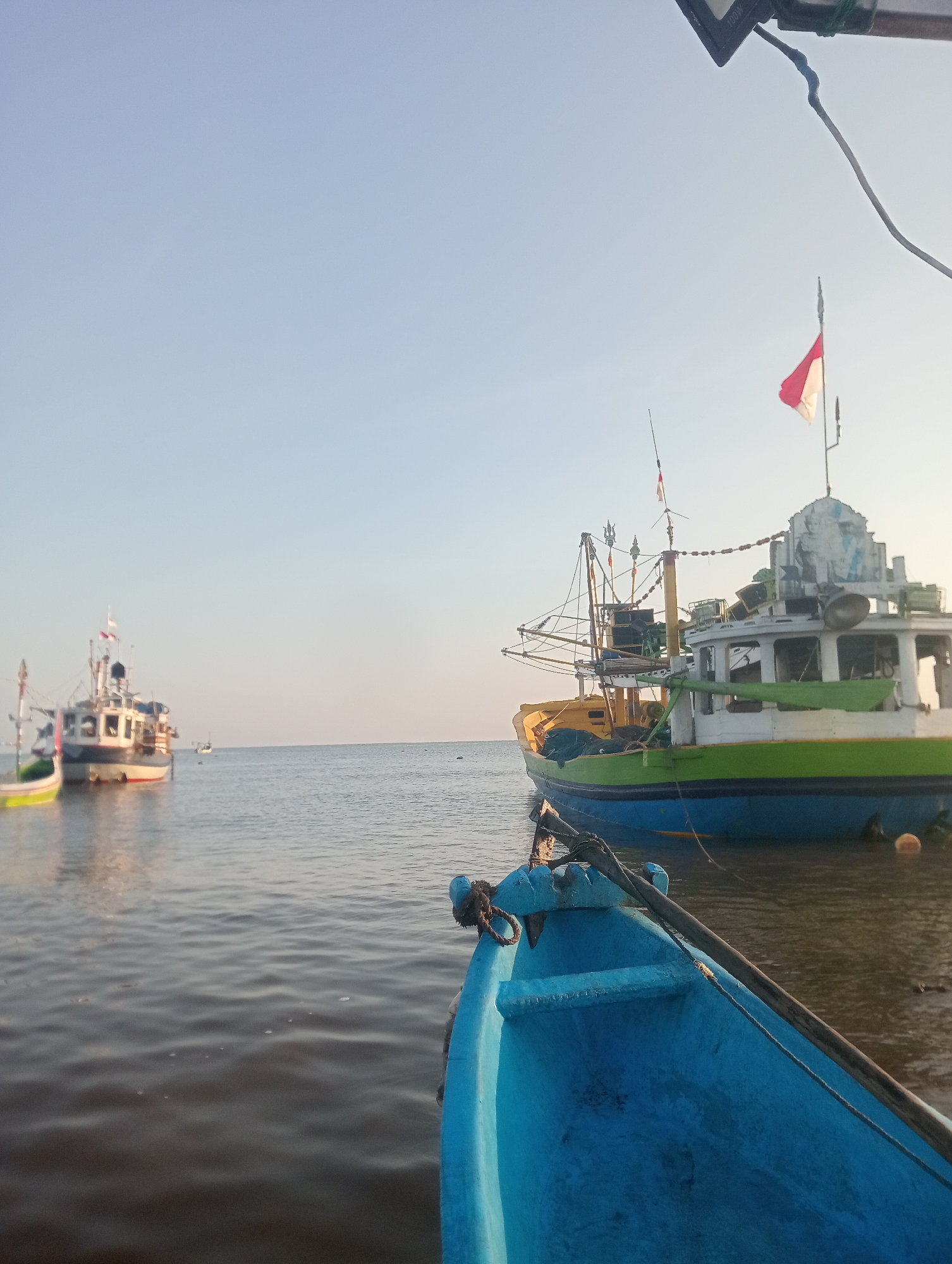 This is a photograph taken from a small blue boat in the foreground, looking out over the water toward several larger fishing boats anchored offshore. The sky is clear and a pale blue, with the sun low on the horizon, suggesting it is either early morning or late afternoon. The water is calm, reflecting the light and the shapes of the boats.
In the middle ground, a larger, colorful fishing boat with a yellow and green hull is prominently featured. It has a white superstructure adorned with what appears to be decorative blue and white imagery. A small Indonesian flag, red and white, flies from its mast. This boat is equipped with fishing gear, including nets and rigging. Behind it, further to the left, is another fishing boat with a white and blue hull, also appearing to be at anchor. In the distance, more boats are visible, scattered across the water, and the horizon line separates the sea from the sky.
The foreground boat, from which the photo is taken, is partially visible, showing its bright blue interior and a rough mooring rope. A wooden oar is also present. The scene is serene and depicts a typical harbor or anchorage scene for fishing vessels.
Based on visual cues such as the Indonesian flag, the style of the fishing boats, and the clear sky typical of tropical regions, this image is likely located in a coastal area of Indonesia. Besuki, a port town in East Java, Indonesia, is known for its fishing activities and coastal landscape, making it a plausible location for this scene.