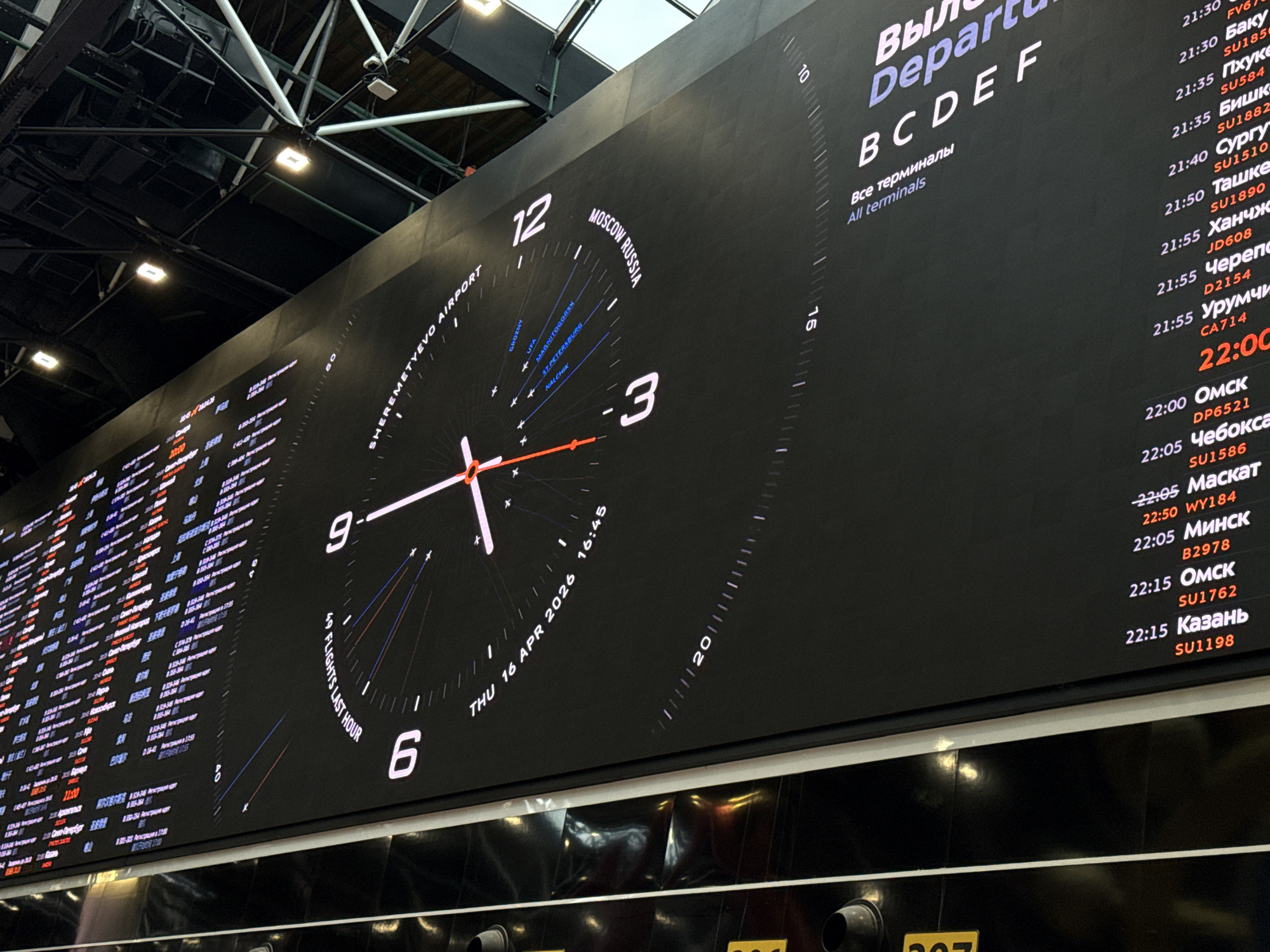 A wide-angle, low-perspective shot captures a large, modern digital departure board inside an airport terminal. The setting is indoors, with a dark ceiling structure featuring exposed beams and bright rectangular lights illuminating the space. No people are visible.
The oversized display features a prominent, minimalist analog clock face with white numbers 12, 3, 6, and 9. White and red hands indicate the time is approximately 16:45, explicitly stated below as "THU 16 APR 2026 16:45". Arcing above the clock is the text "SHEREMETYEVO AIRPORT" and "MOSCOW RUSSIA". Radial lines extend from the clock's center, pointing to various Russian cities like GROZNY, UFA, MAGNITOGORSK, ST. PETERSBURG, and NALCHIK. Below the 6, "49 FLIGHTS LAST HOUR" is displayed.
To the right, a detailed list of departures is presented against the dark background, with white and red text for clarity. The heading reads "Вылет / Departures" for "Все терминалы / All terminals B C D E F". Flights are listed chronologically, starting from 21:30 and extending past 22:15. Destinations include Баку (Baku), Пхукет (Phuket), Бишкек (Bishkek), Сургут (Surgut), Ташкент (Tashkent), Ханчжоу (Hangzhou), Череповец (Cherepovets), Урумчи (Urumqi), Омск (Omsk), Чебоксары (Cheboksary), Маскат (Muscat), Минск (Minsk), and Казань (Kazan), each with a corresponding flight number (e.g., SU185, JD608, WY184).
The left section of the board also shows extensive flight information, though it is less distinct due to the angle. The overall scene depicts a functional, informative environment typical of a major airport, specifically Sheremetyevo Airport in Khimki, Russia.